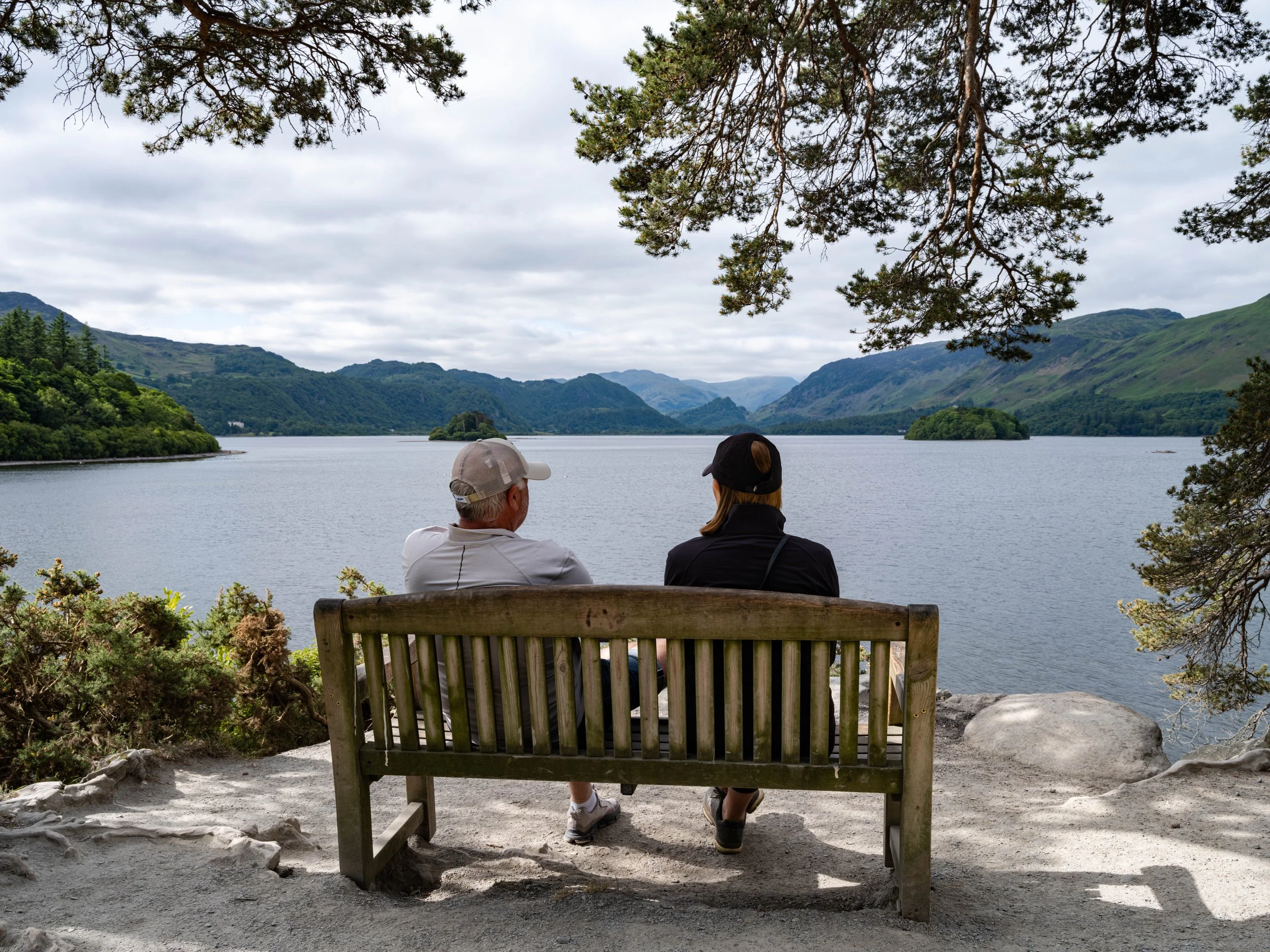 Two people sitting on a wooden bench overlooking a lake with green mountains in the background, under a partly cloudy sky.