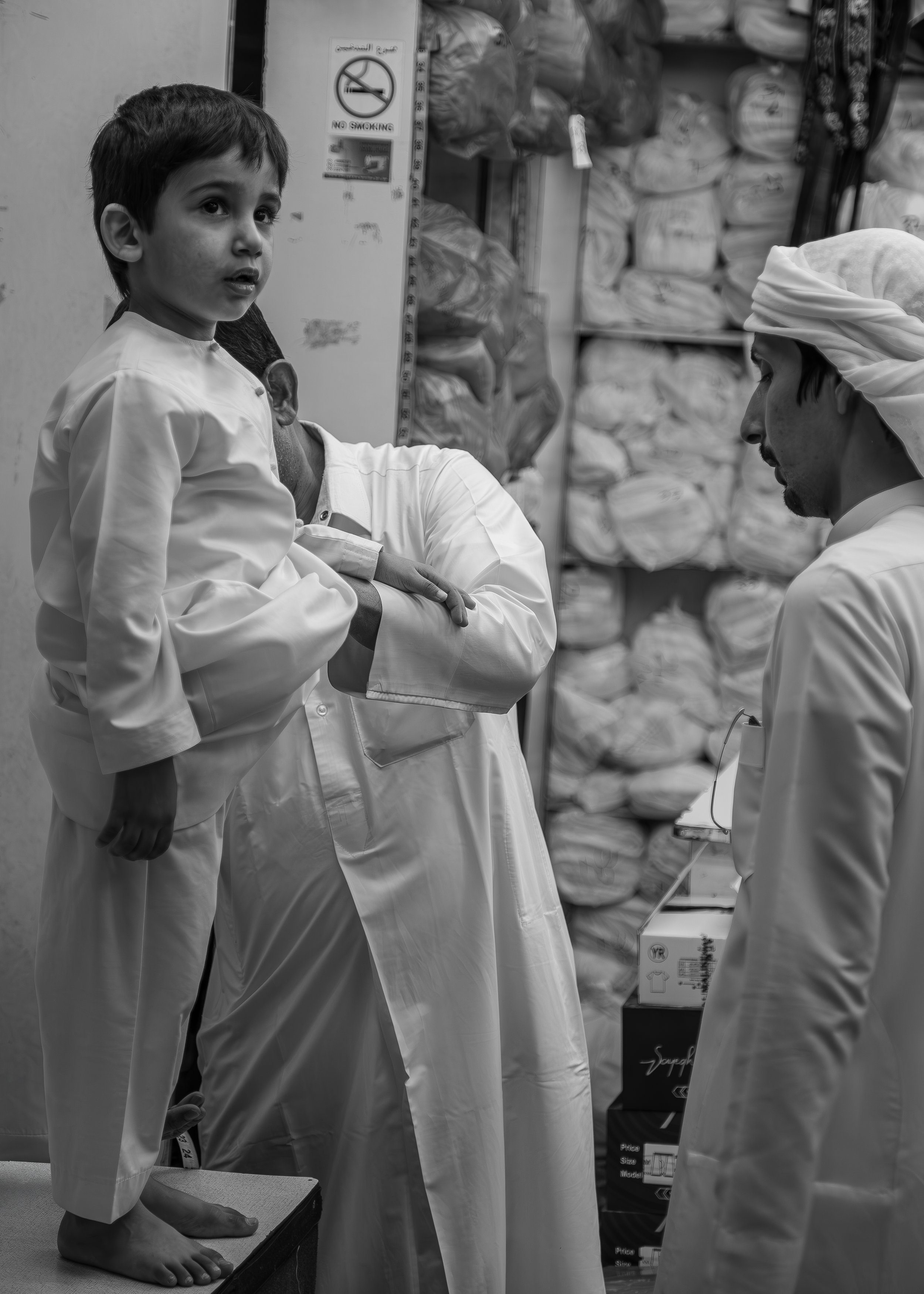 A young boy standing on a platform in a store surrounded by men in traditional Middle Eastern attire, with shelves and packaged goods in the background, in a black-and-white photograph.
