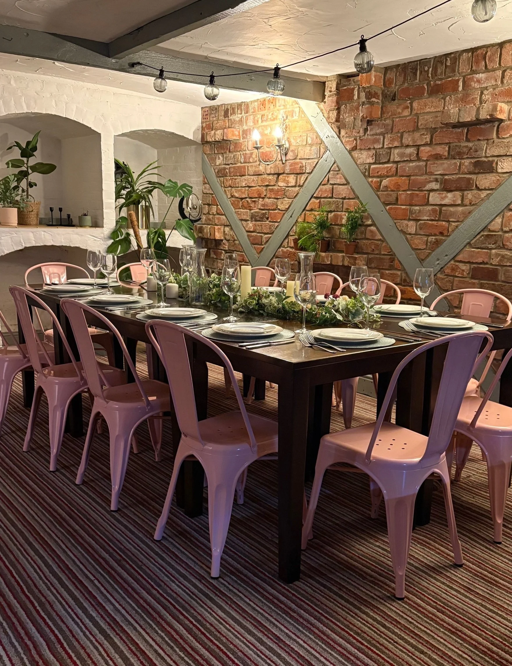 A long dining table set for a formal meal with white plates, wine glasses, and pink wine bottles, decorated with vases of purple and pink flowers in front of a brick wall with candles and greenery. Private chef, enquire for celebration menus. 