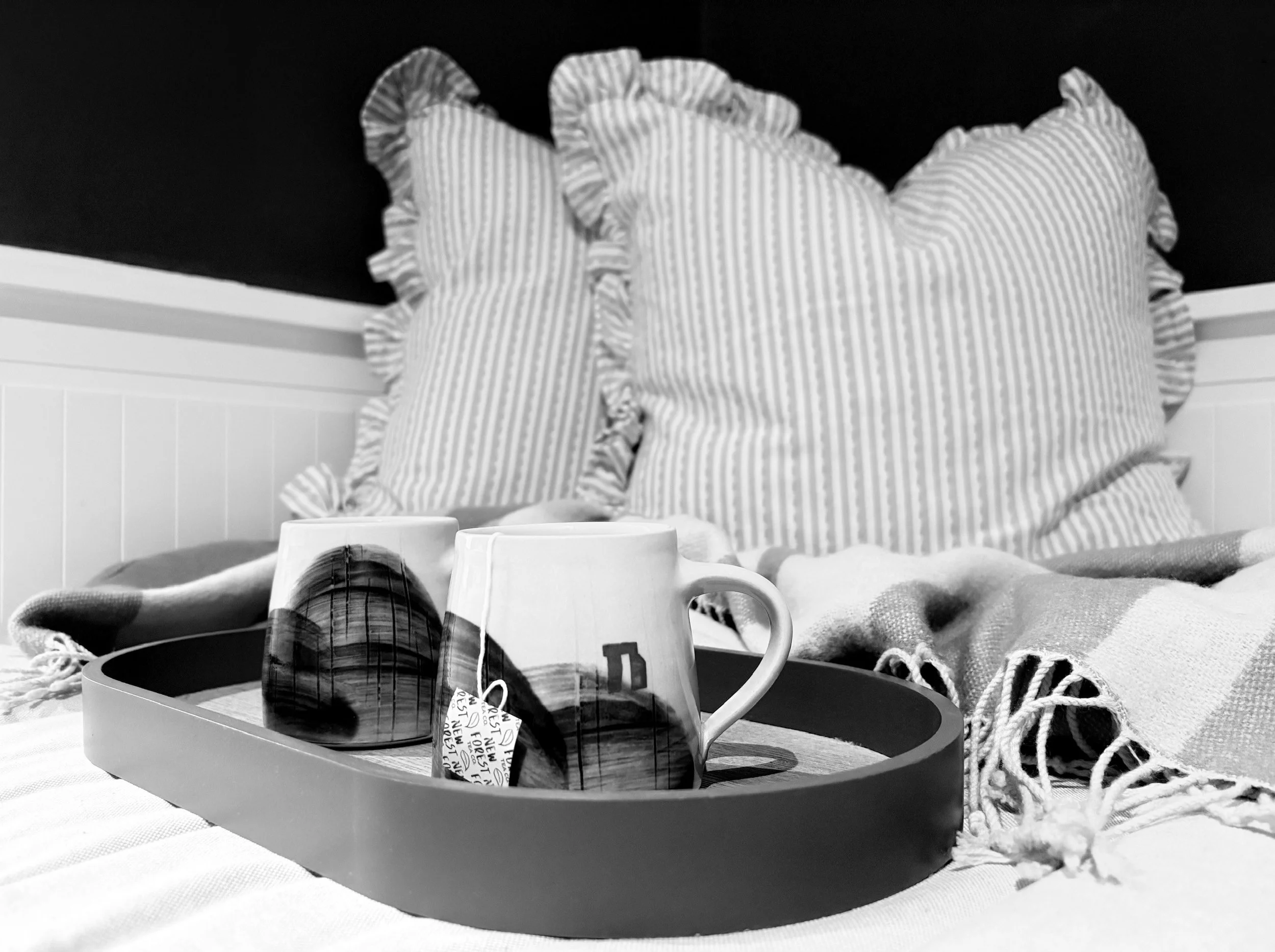 A tray with two coffee mugs featuring an image of a domed building and a bicycle, placed on a bed with striped ruffled pillows and a blanket.