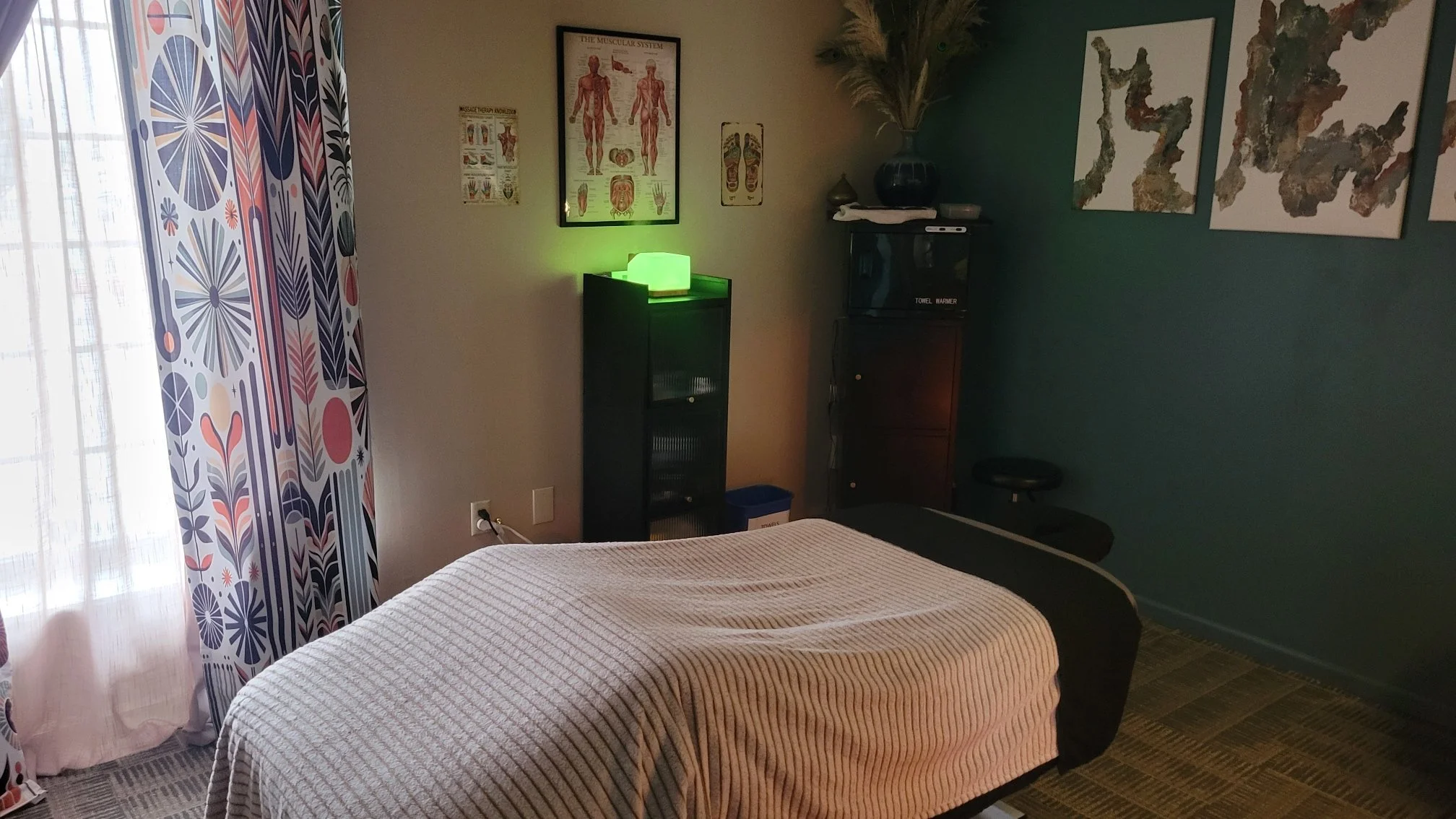 A massage therapy room with a massage bed covered in a beige textured sheet, colorful patterned curtains on a window to the left, framed anatomical posters on the beige wall, a black cabinet with a glowing green light on top, and a dark green wall with abstract art pieces.