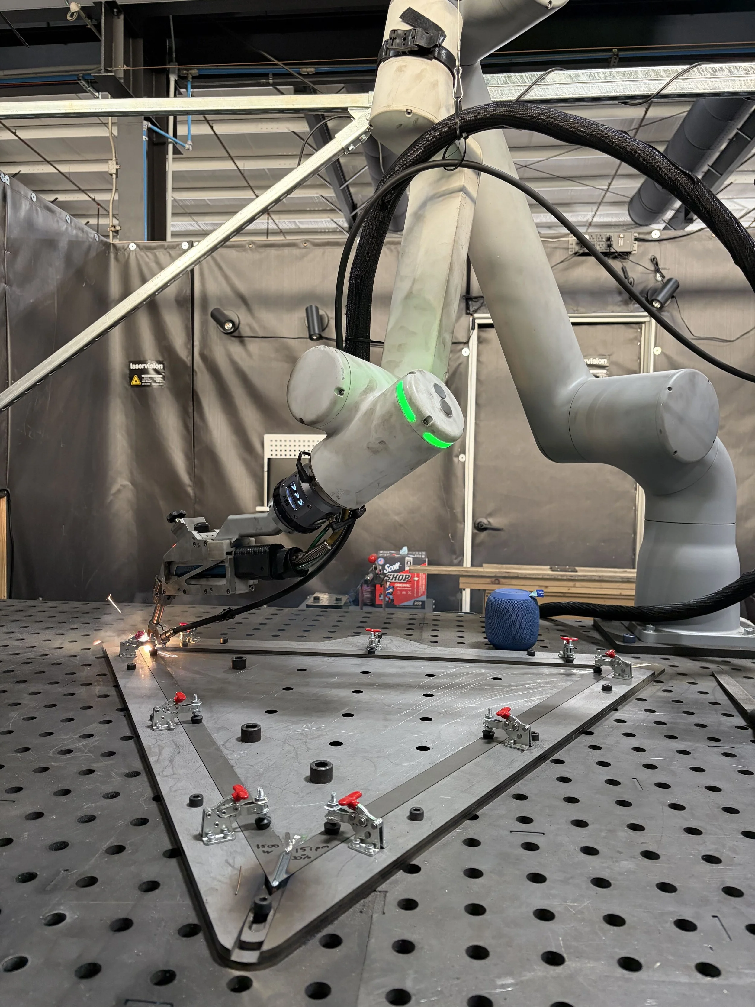 Industrial robotic arm welding a metal component on a perforated workbench inside a fabrication facility.