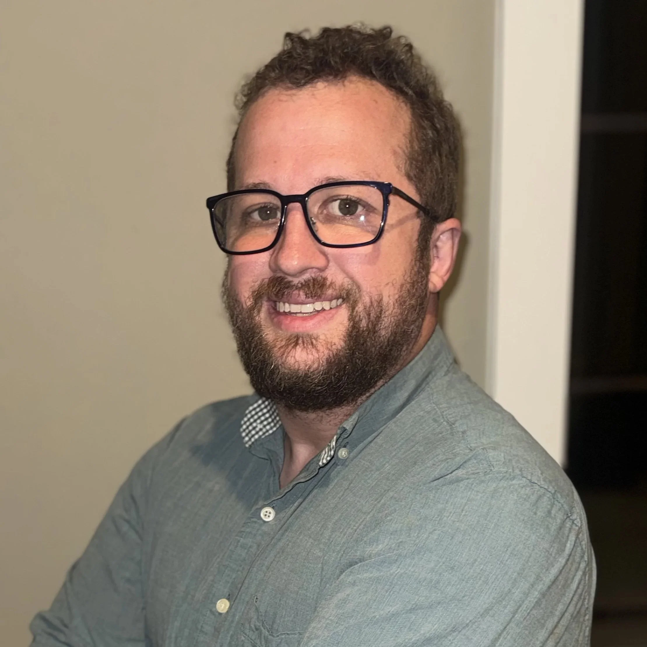 A smiling man with glasses and a beard wearing a gray shirt, standing indoors in front of a beige wall.
