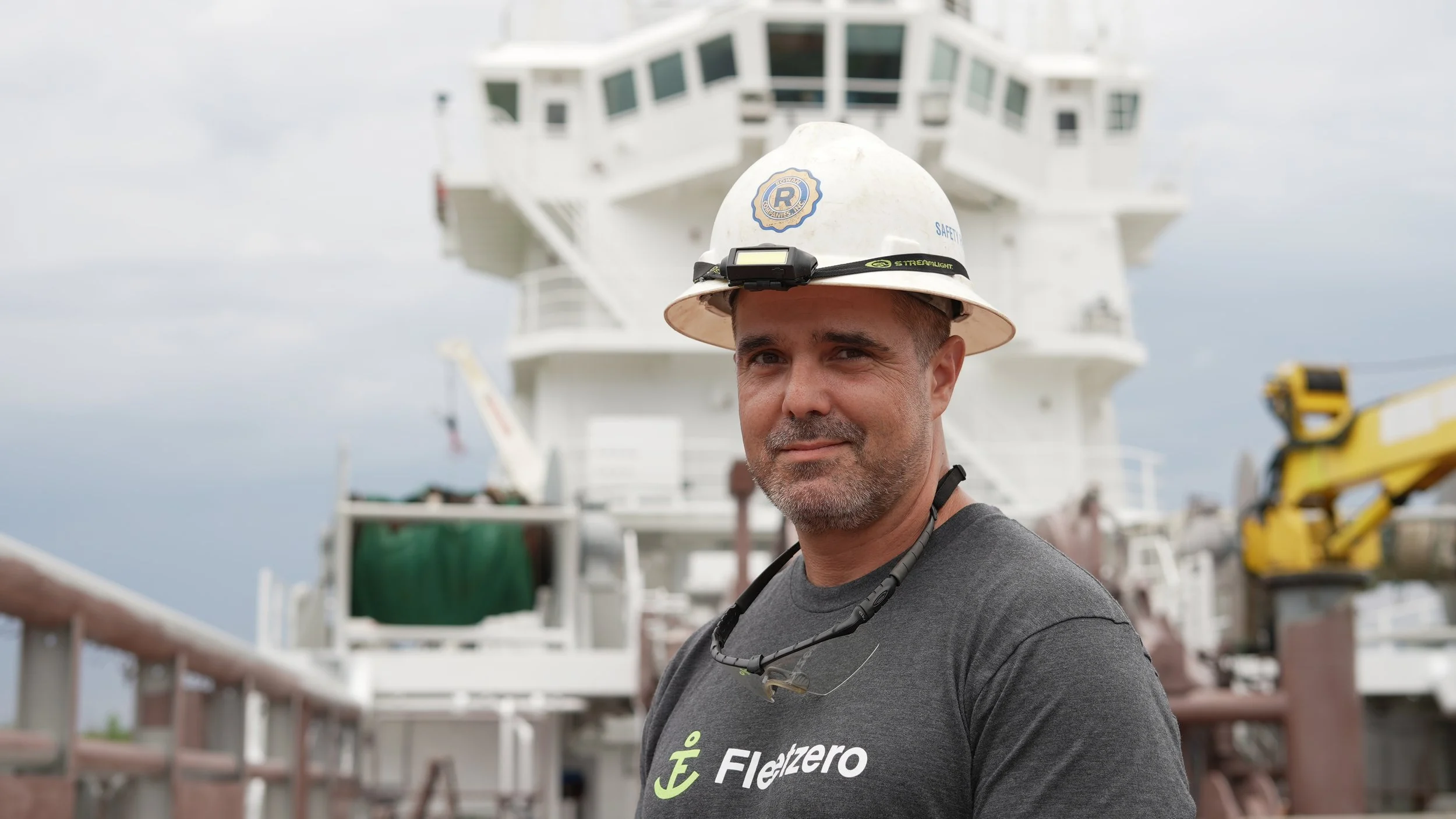Man wearing a hard hat and headlamp aboard a ship, with a yellow crane in the background