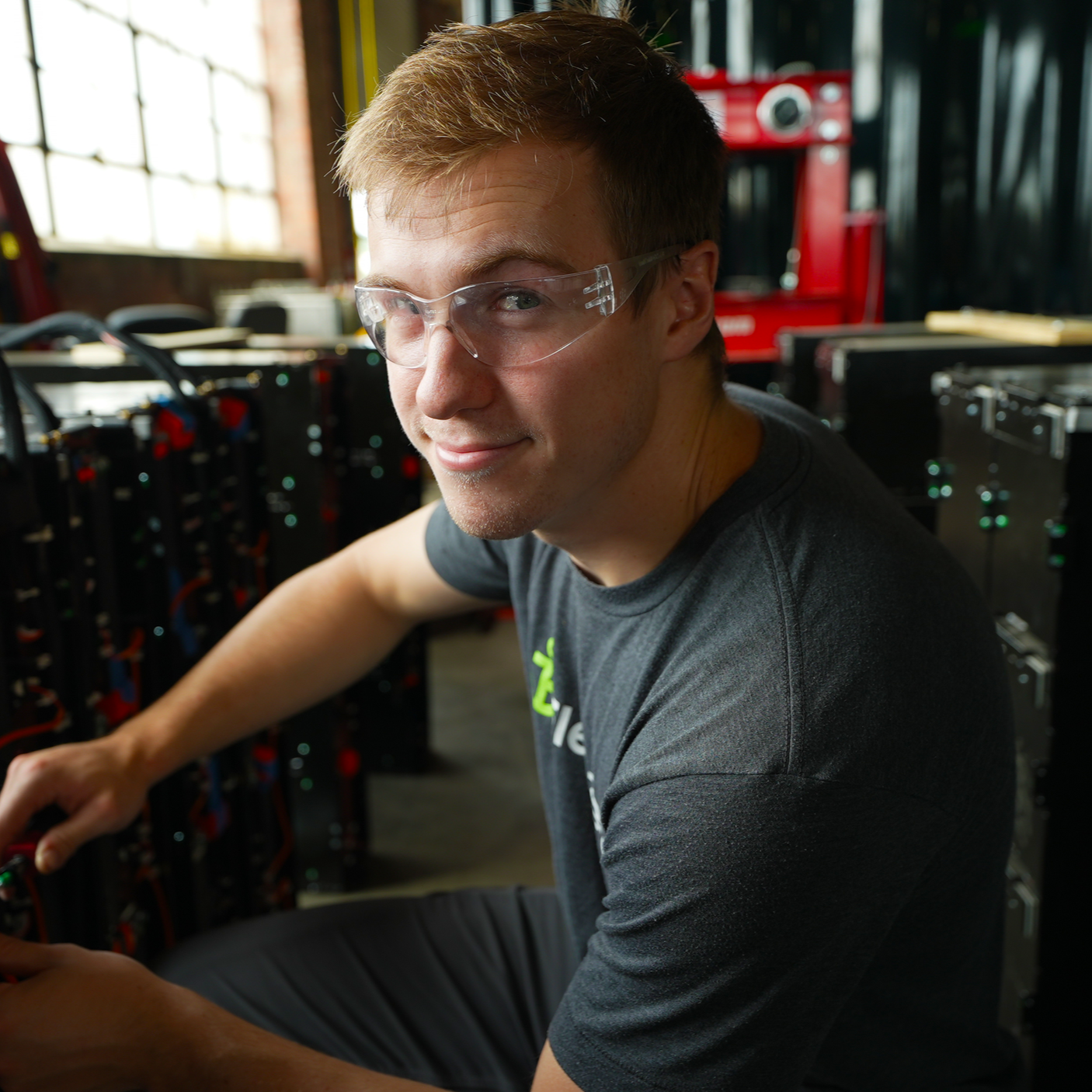 A man with light brown hair wearing safety glasses working with electronic equipment in a workshop or data center, with server racks and tools around him.