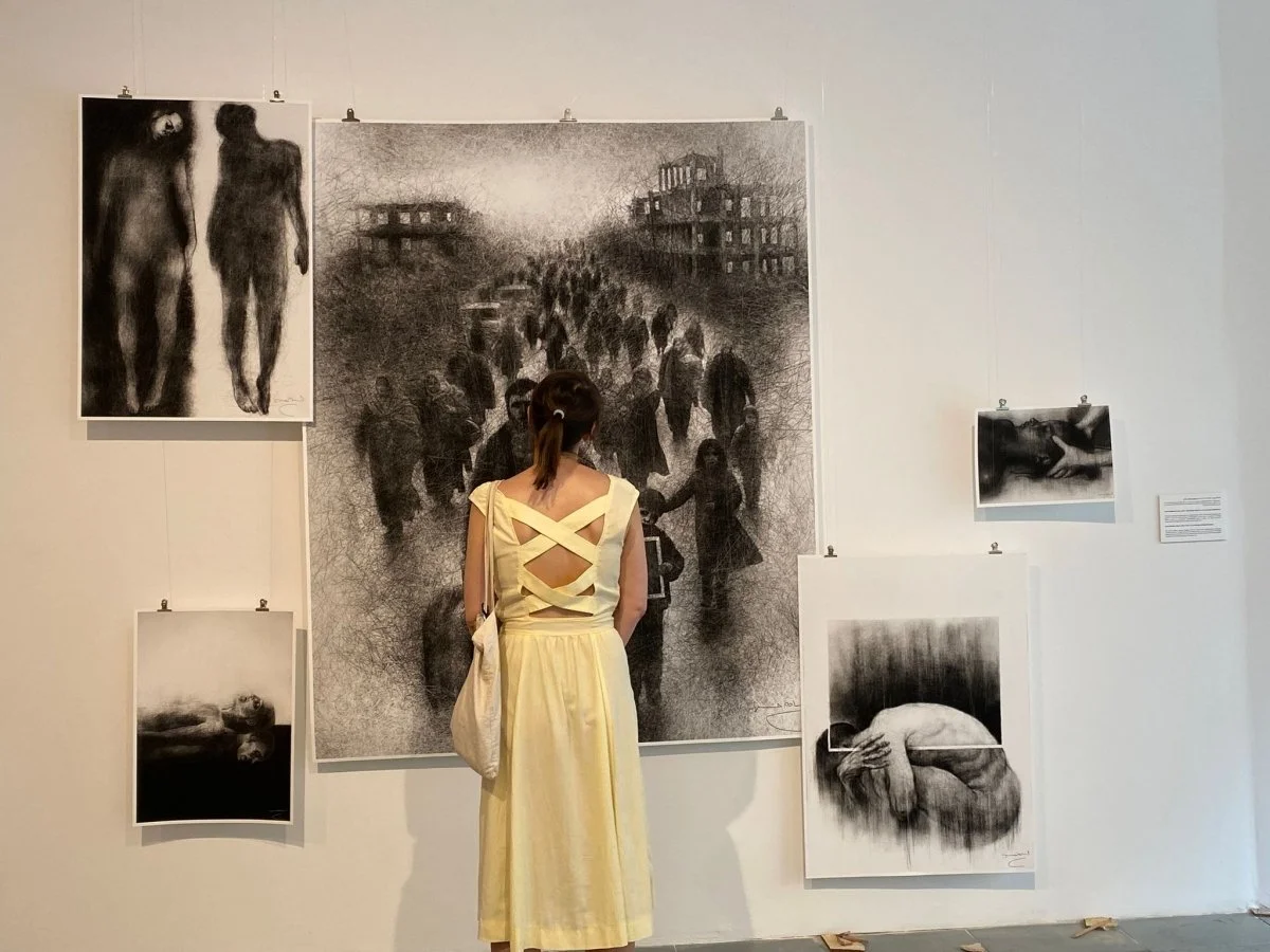 A woman in a yellow dress stands in an art gallery, observing large black-and-white drawings on the wall, including a depiction of a crowd, two figures walking, a reclining figure, and a hand.