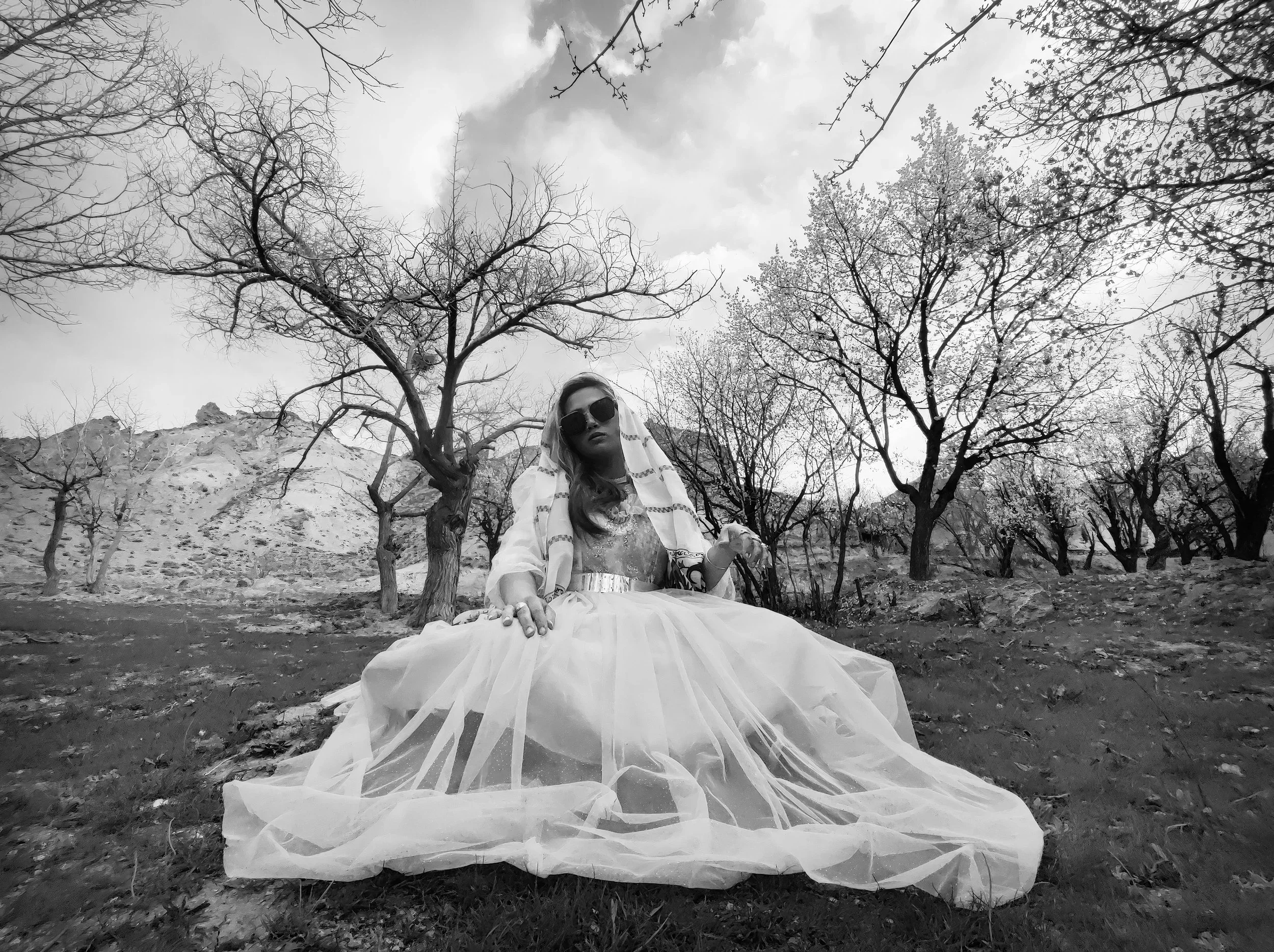 A woman wearing large sunglasses, a intricately patterned dress, and a sheer layered skirt with a metallic belt, sitting on the ground in a leafless trees landscape, with hills in the background. The image is in black and white.