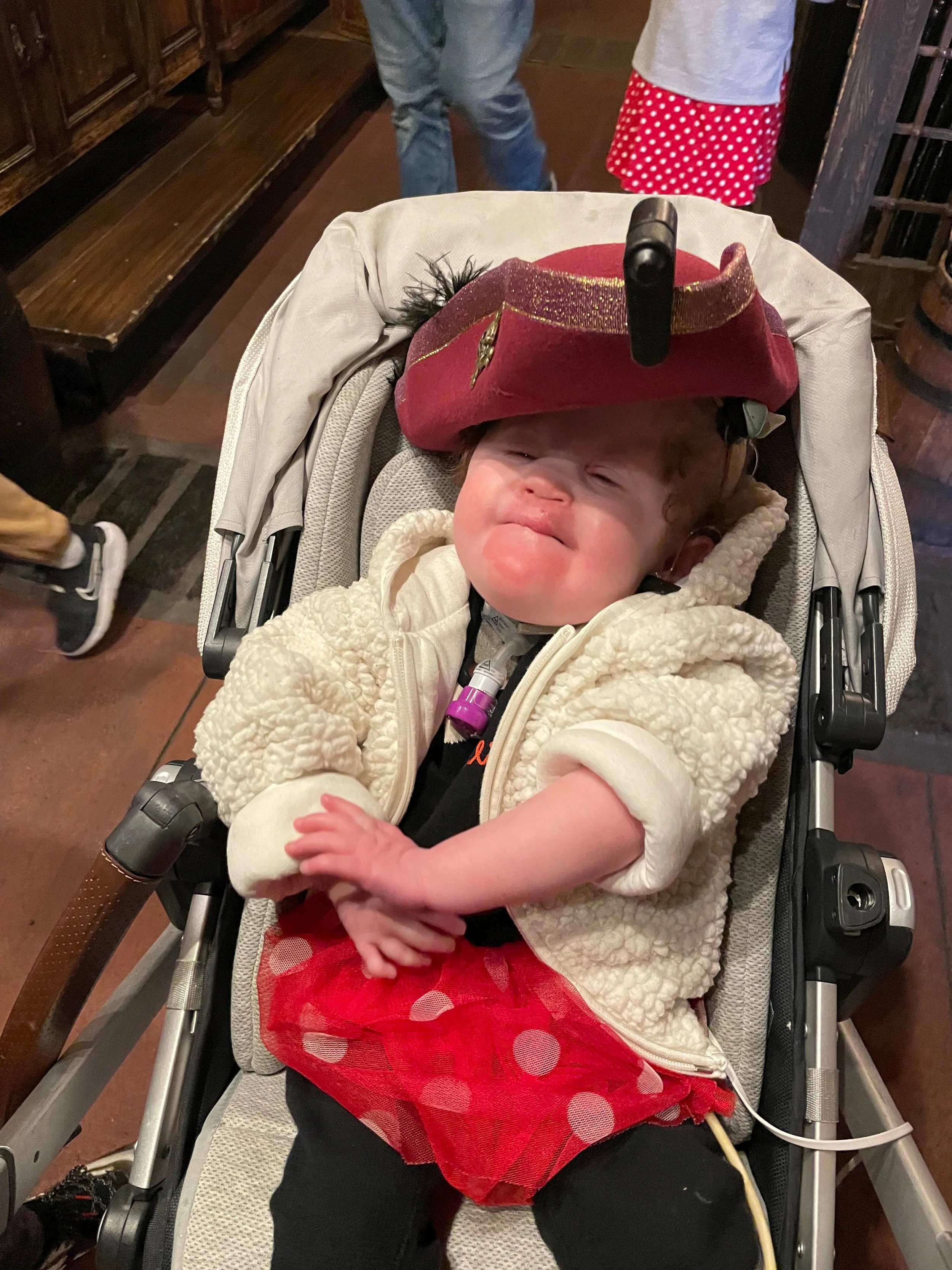 A young girl with a sad or upset expression sitting in a stroller wearing a red and white polka dot tutu, a cream-colored fleece jacket, and a red topper resembling a pirate hat.