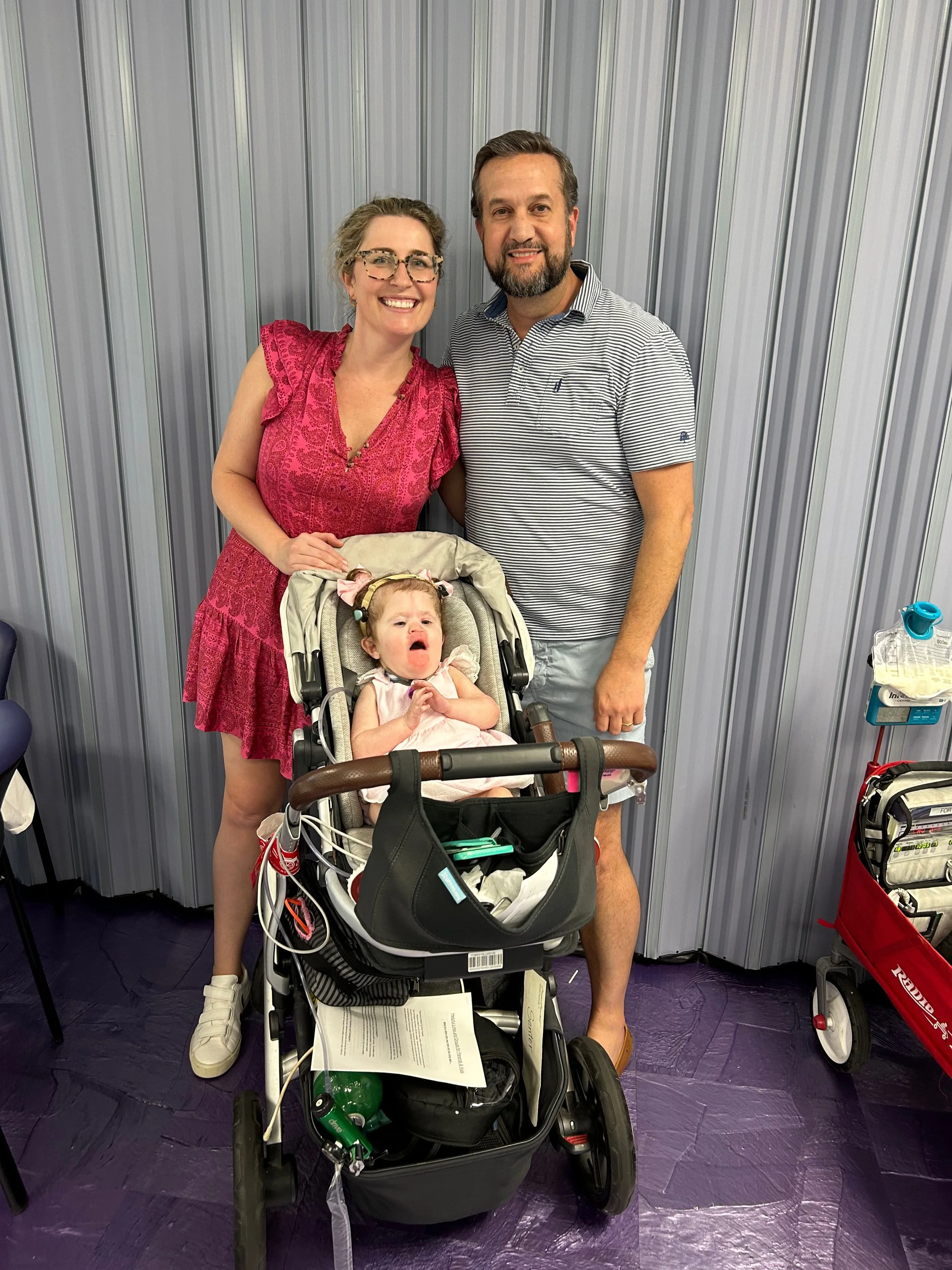 A family of four standing together at a hospital or clinic. A woman in a pink dress, a man in a striped polo shirt, and a girl in a stroller with medical equipment. The girl appears to be unwell, with a concerned expression.