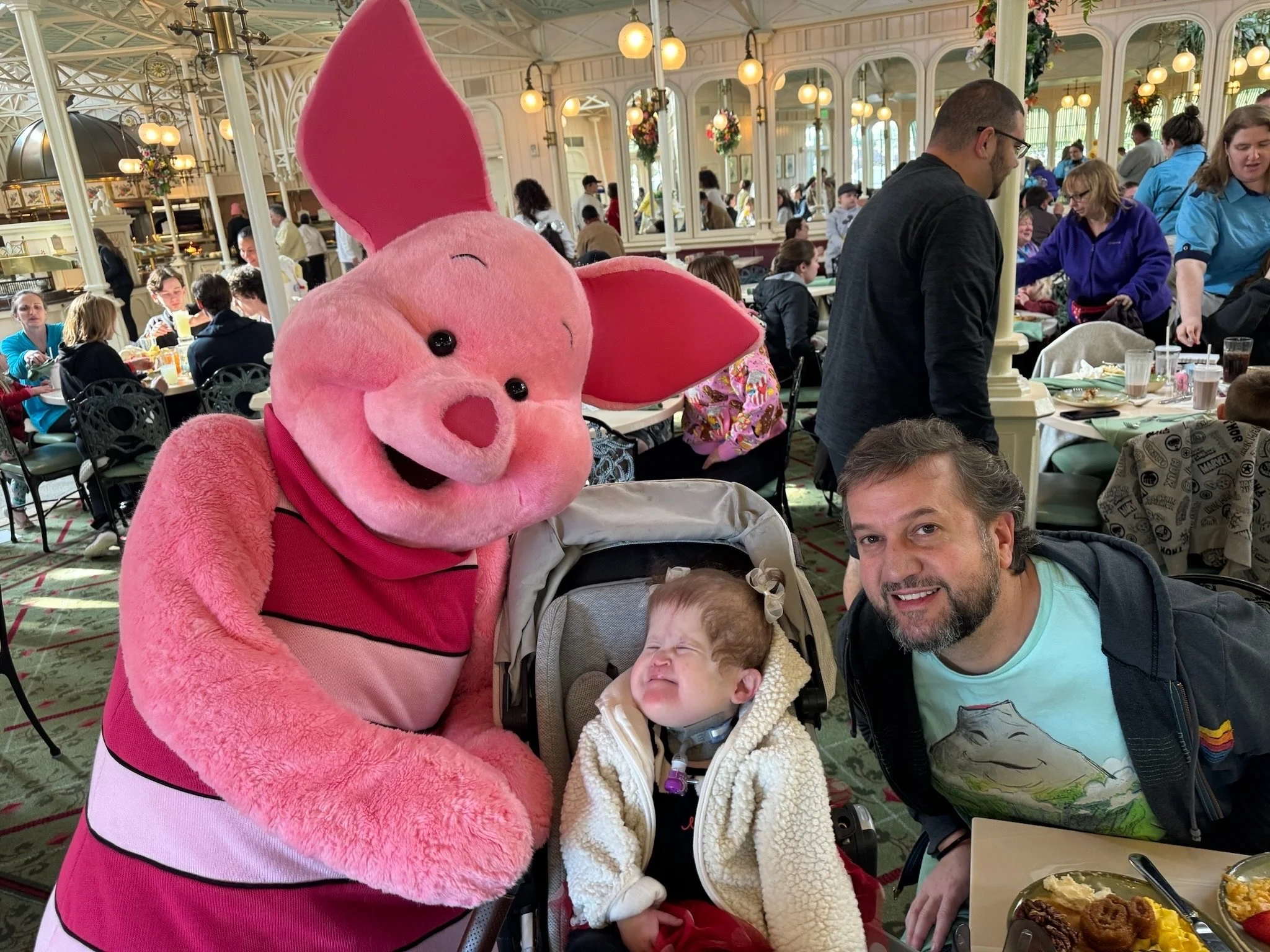 A themed character dressed as Piglet from Winnie the Pooh, standing next to a woman and a young child sitting in a stroller at a busy restaurant or cafe with many people dining at tables.