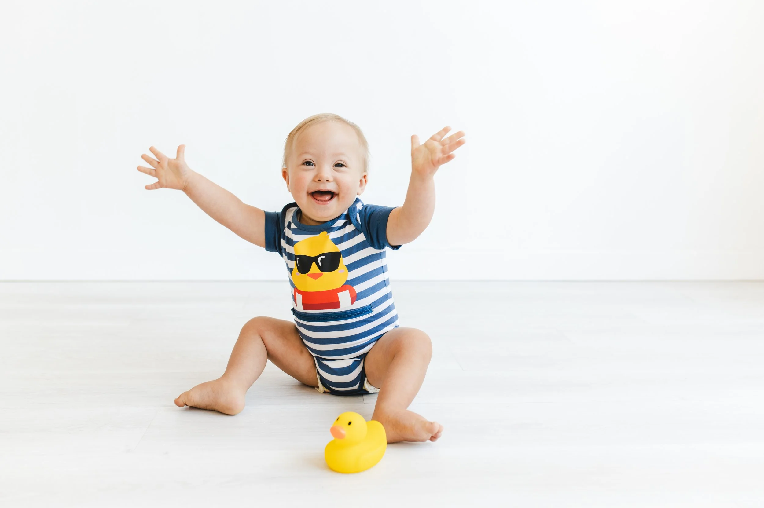 A happy baby sitting on a white floor with a yellow rubber duck, wearing a striped blue and white t-shirt with a yellow thumbs up cartoon with sunglasses, arms outstretched.