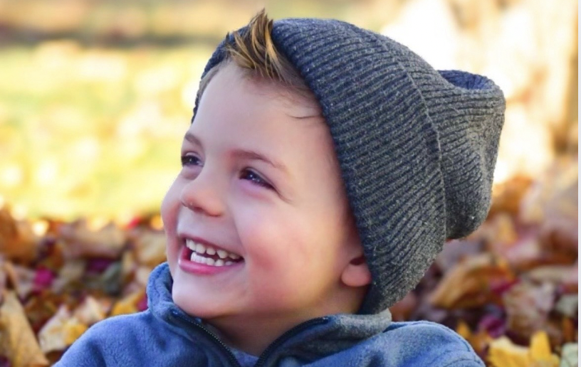 A young boy smiling outdoors in a setting with fallen autumn leaves, wearing a dark gray beanie and a blue jacket.