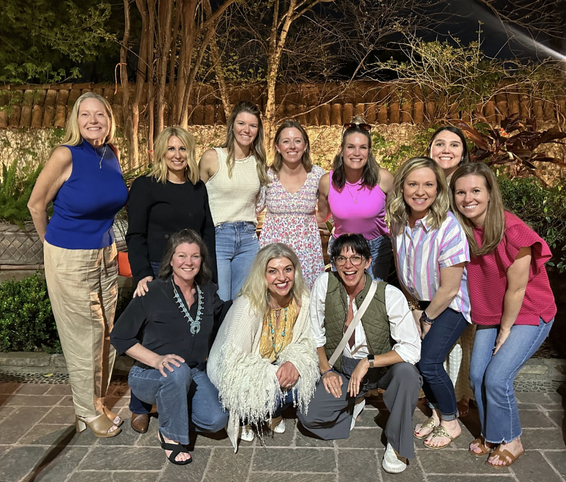 A group of twelve women gathered outdoors at night, smiling and posing for a photo, with trees and a wooden fence in the background.