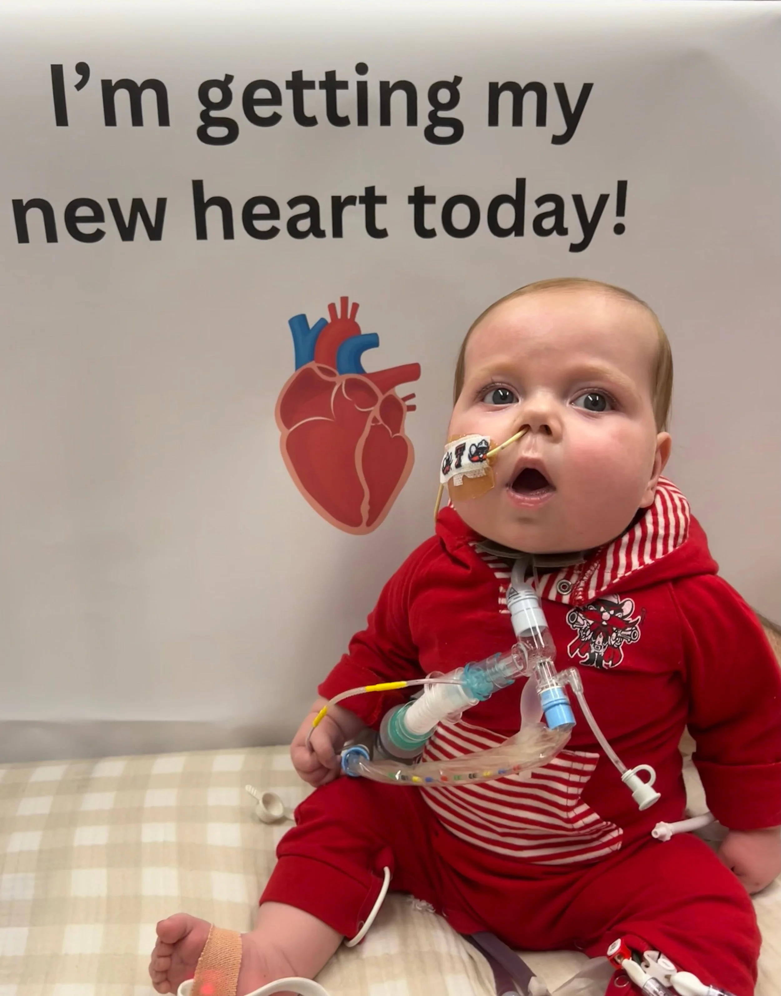 A baby in a red hoodie and red pants sitting on a hospital bed with a heart-shaped illustration and the text 'I'm getting my new heart today!' in the background. The baby has medical equipment attached to their chest and a nasal tube, looking surprised.