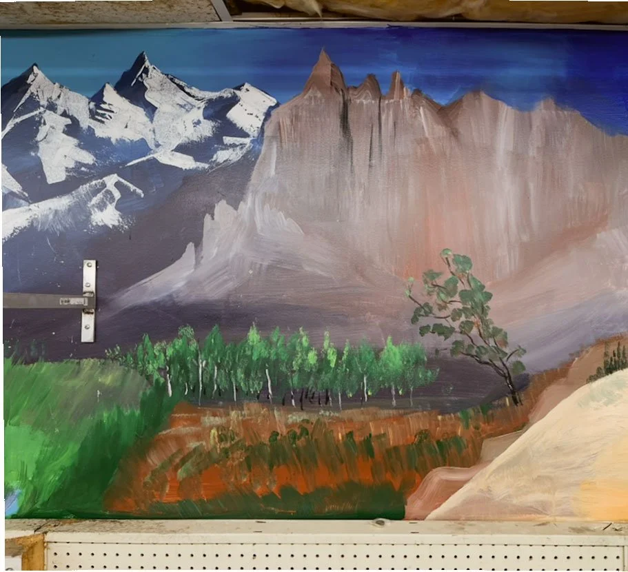 snow capped mountains on left side leading into brown mountains with small forest in foreground and sand dune in lower right corner