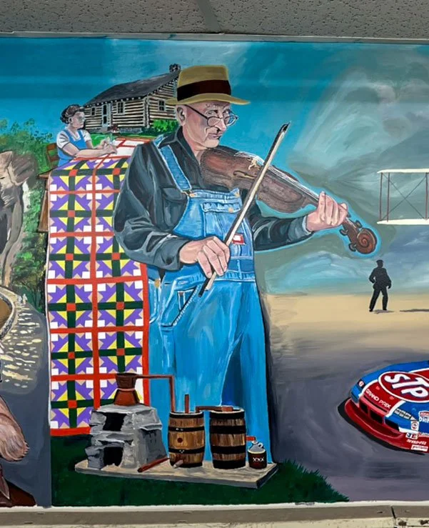 woman sewing quilt in left background, man playing fiddle in center. Moonshine still in front of him. front of racecar to his right, and part of Wright Brother's plane can be seen back right