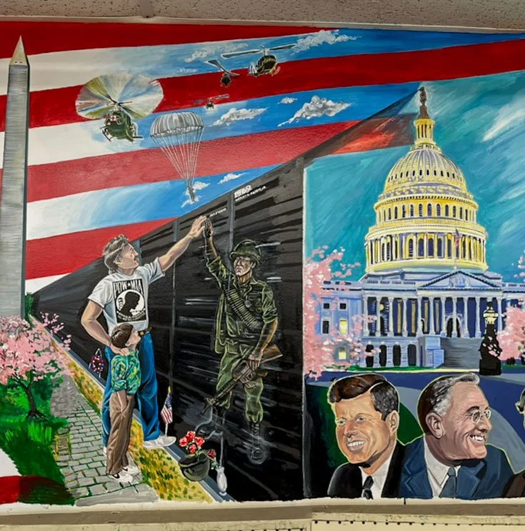 Washington monument in front of American Flag background, cherry blossoms in front of it. Vietnam War Memorial Wall stretches back next to it, with man reaching up to touch wall, silhouette of soldier in wall reaches up to touch his hand. Man's other