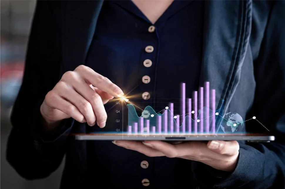 Person in dark jacket and shirt holding a tablet with digital charts and graphs, including bar and line graphs, and a globe icon.