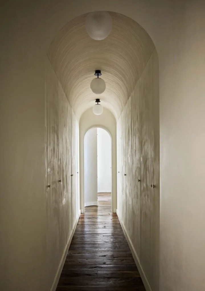 A narrow hallway with light-colored walls, arched ceiling, three spherical ceiling lights, and a wooden floor, leading to a bright doorway at the end.