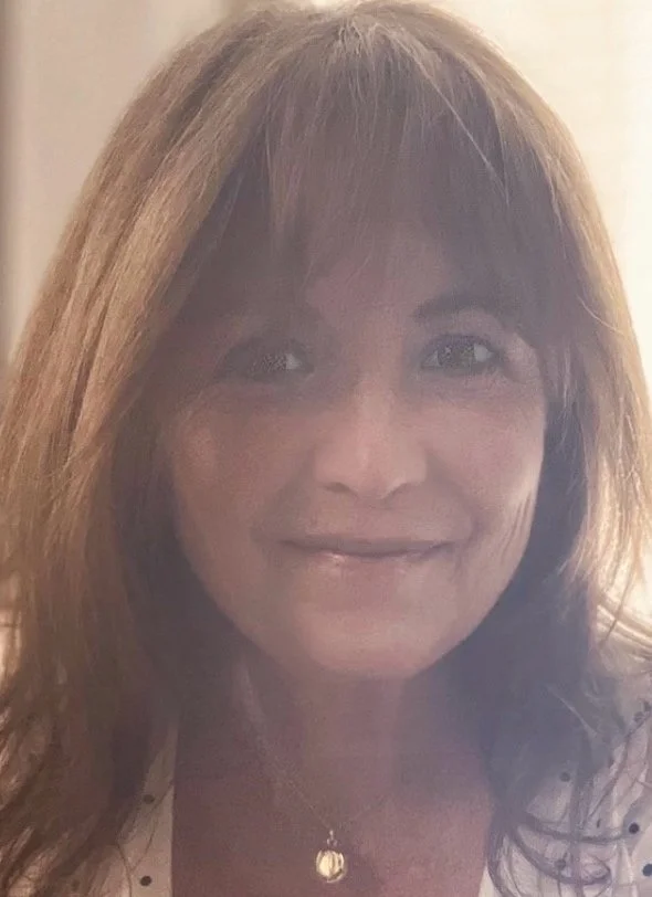 Close-up of a woman with shoulder-length light brown hair smiling, wearing a white top with polka dots and a small pendant necklace.