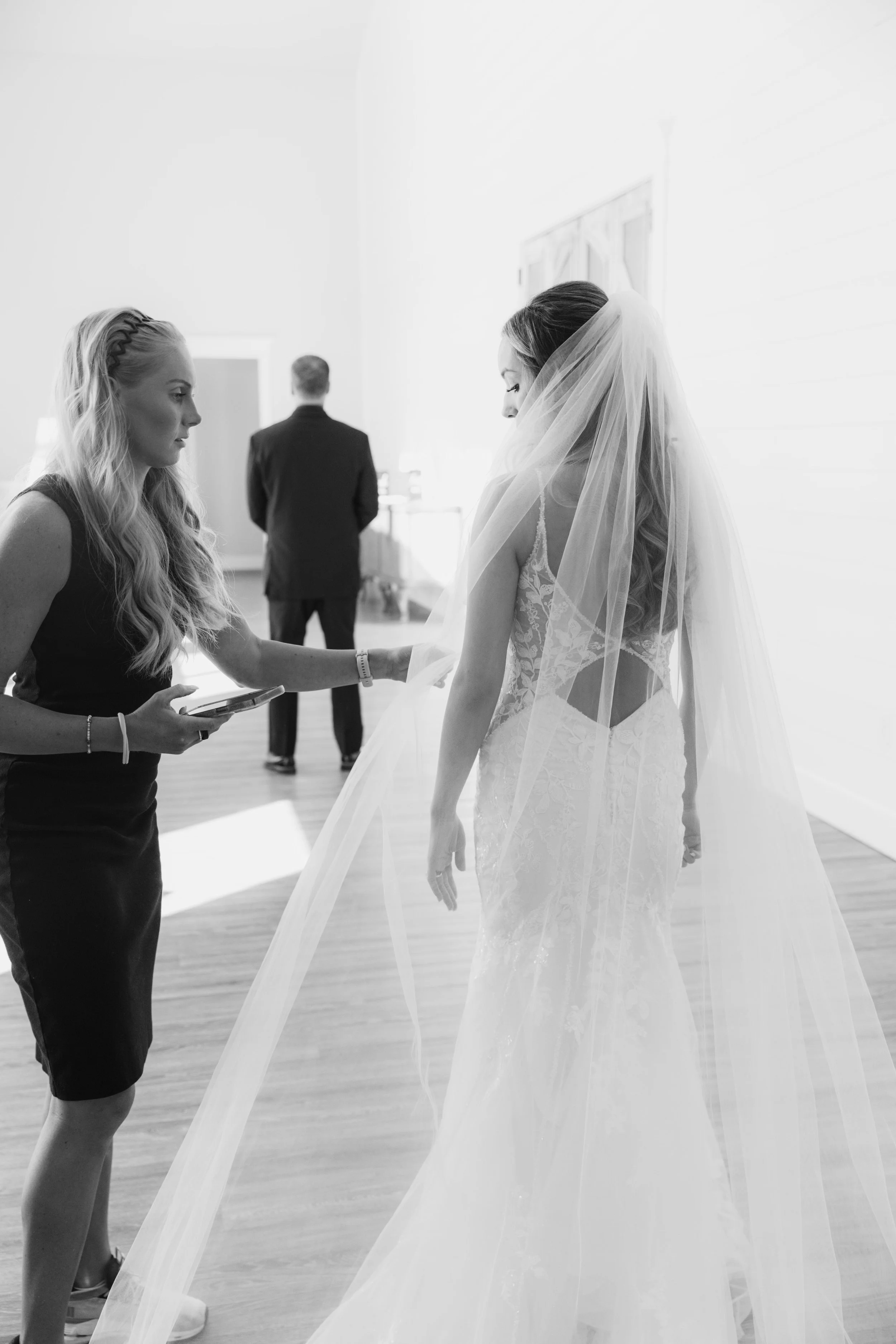 A bride in a wedding dress with a veil standing in a room, with a woman assisting her and a man in a suit in the background.