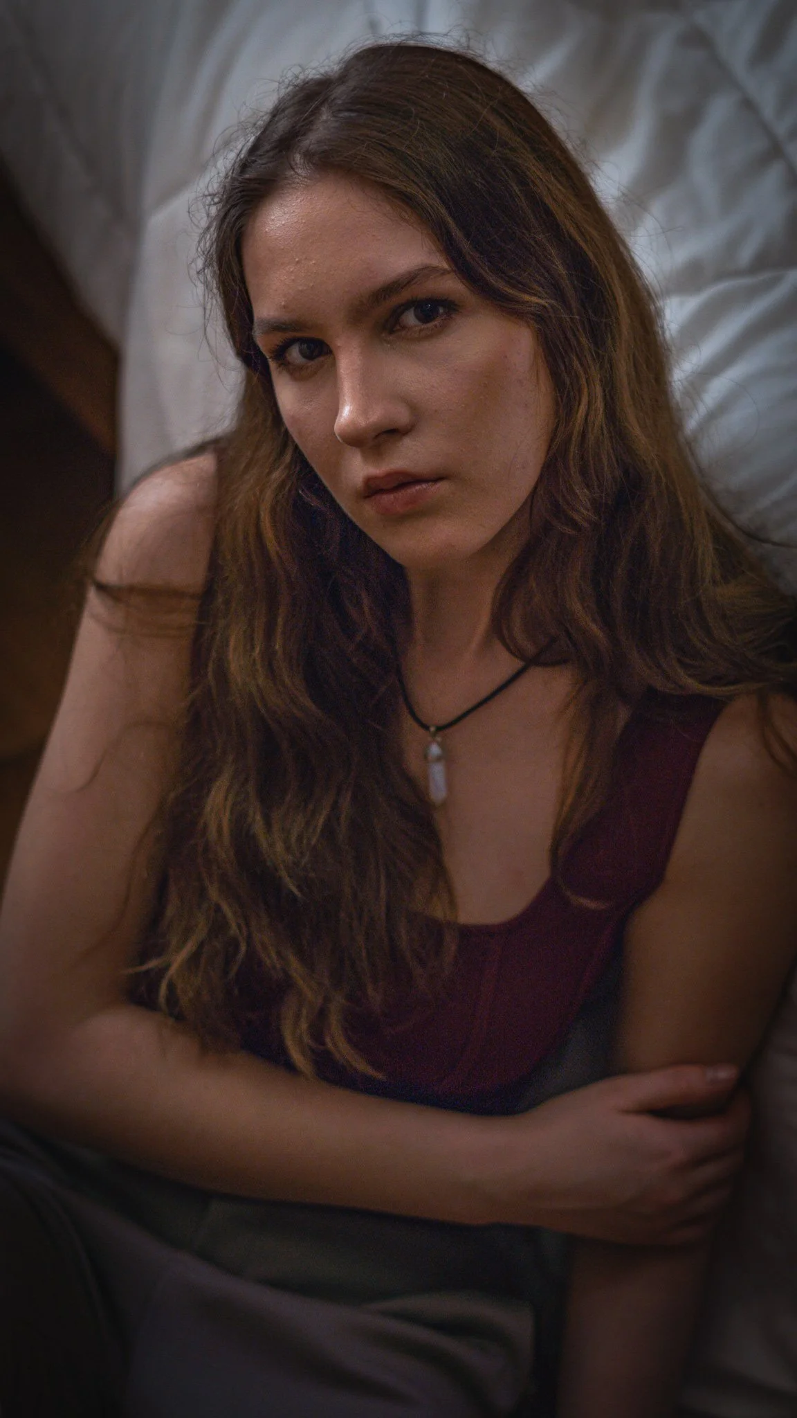 A woman with long, wavy brown hair and light skin lying on her side on a bed, looking at the camera with a serious expression, wearing a dark red top and a necklace with a pendant.