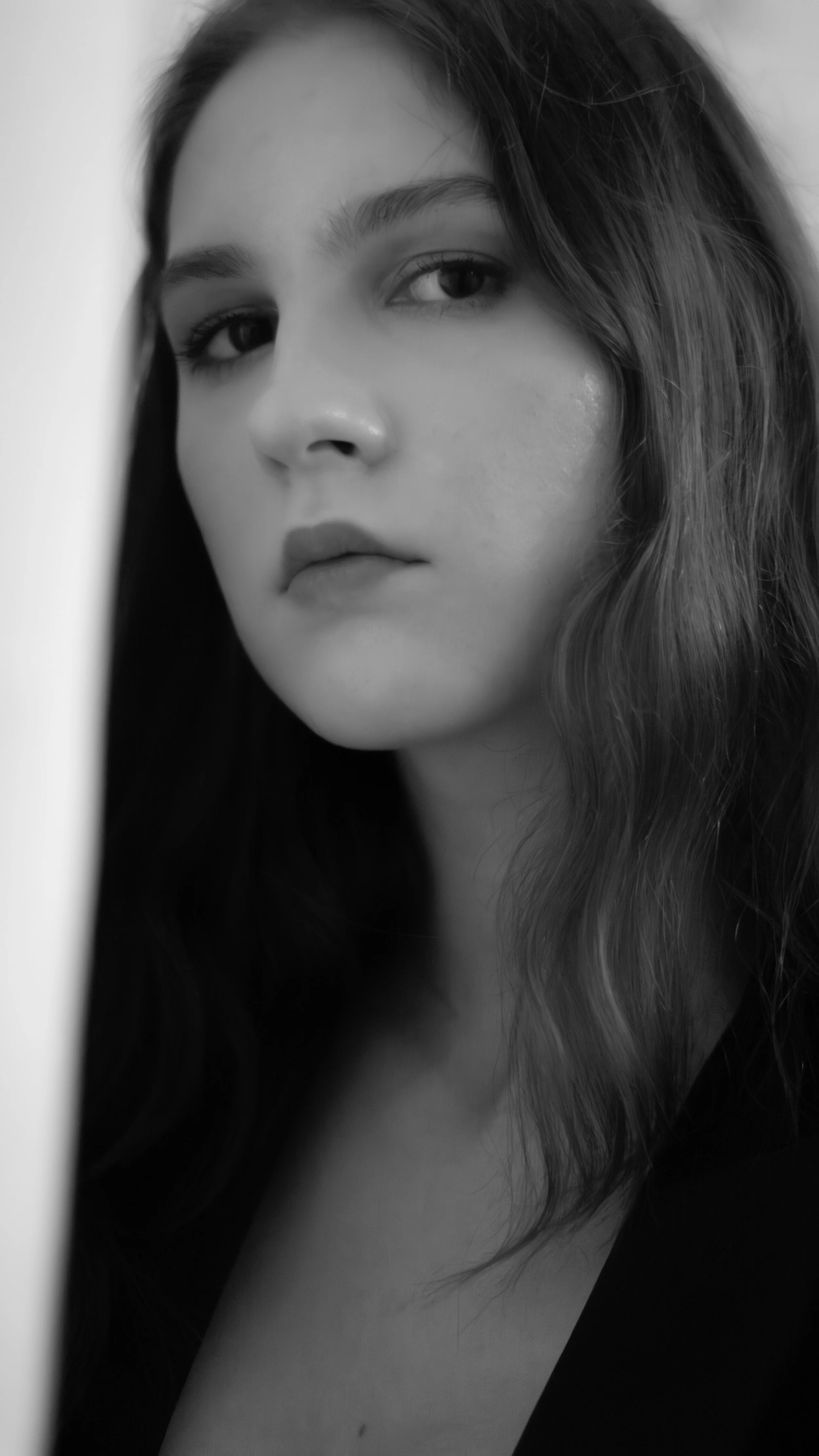 Close-up black and white photograph of a young woman's face, with long wavy hair partially covering her face, looking directly at the camera with a neutral expression.
