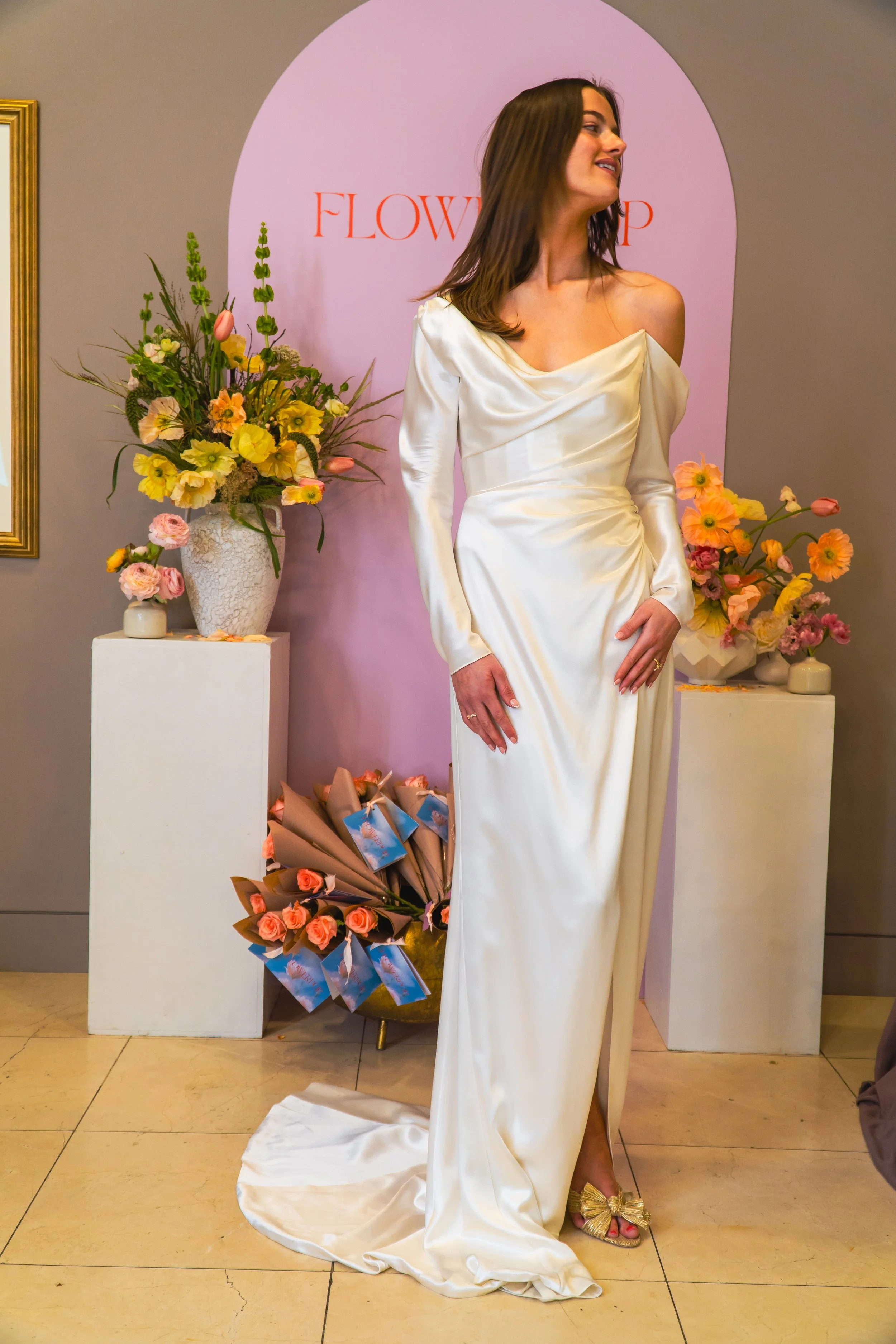 A woman in an off-shoulder ivory satin wedding dress posing indoors near floral arrangements and a pink backdrop with the word 'FLOWERS' on a pink arch.