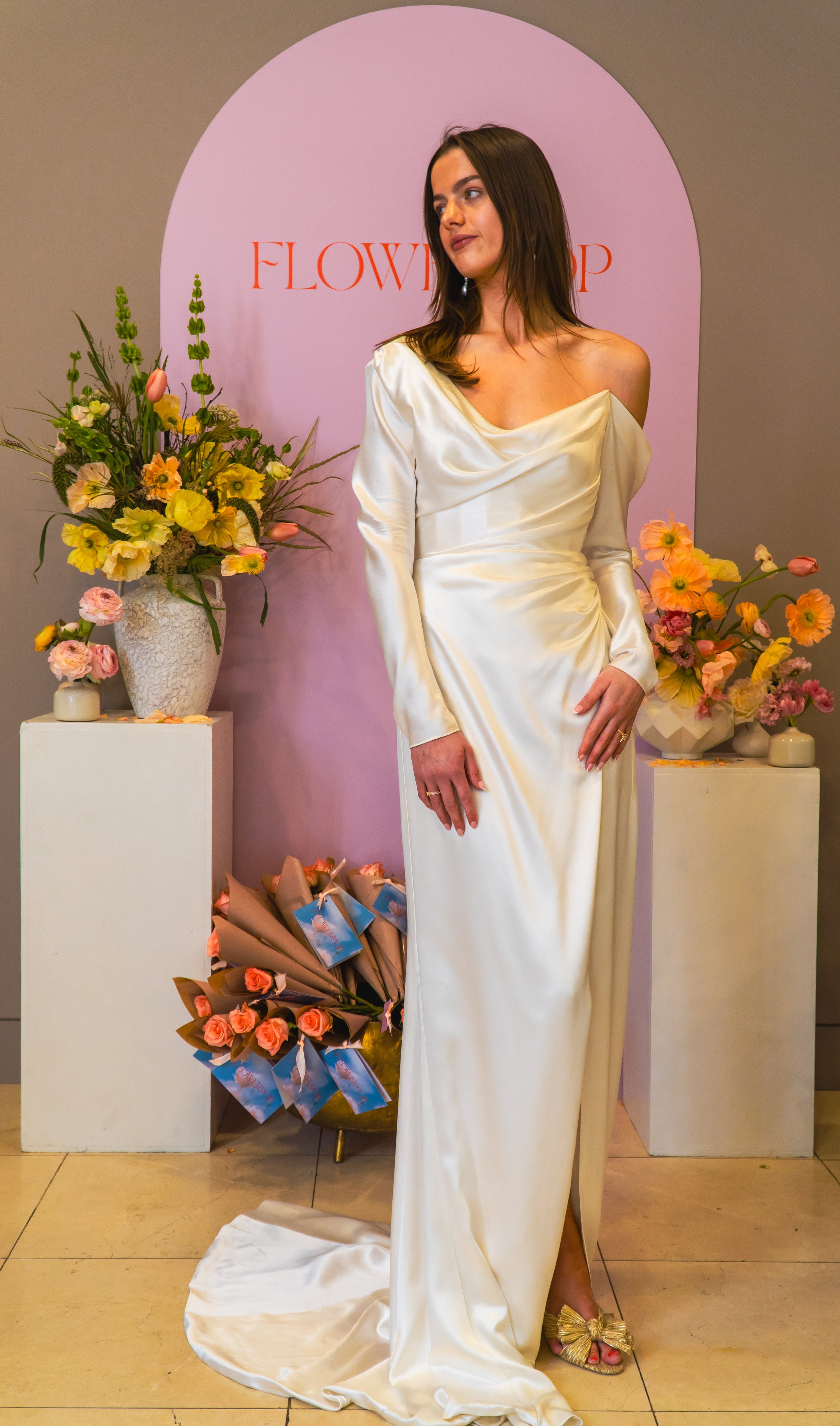 A woman in an off-shoulder, satin cream-colored dress standing in front of a floral display with pink, yellow, and peach flowers, and a pink backdrop with the word 'FLOWER' on it.