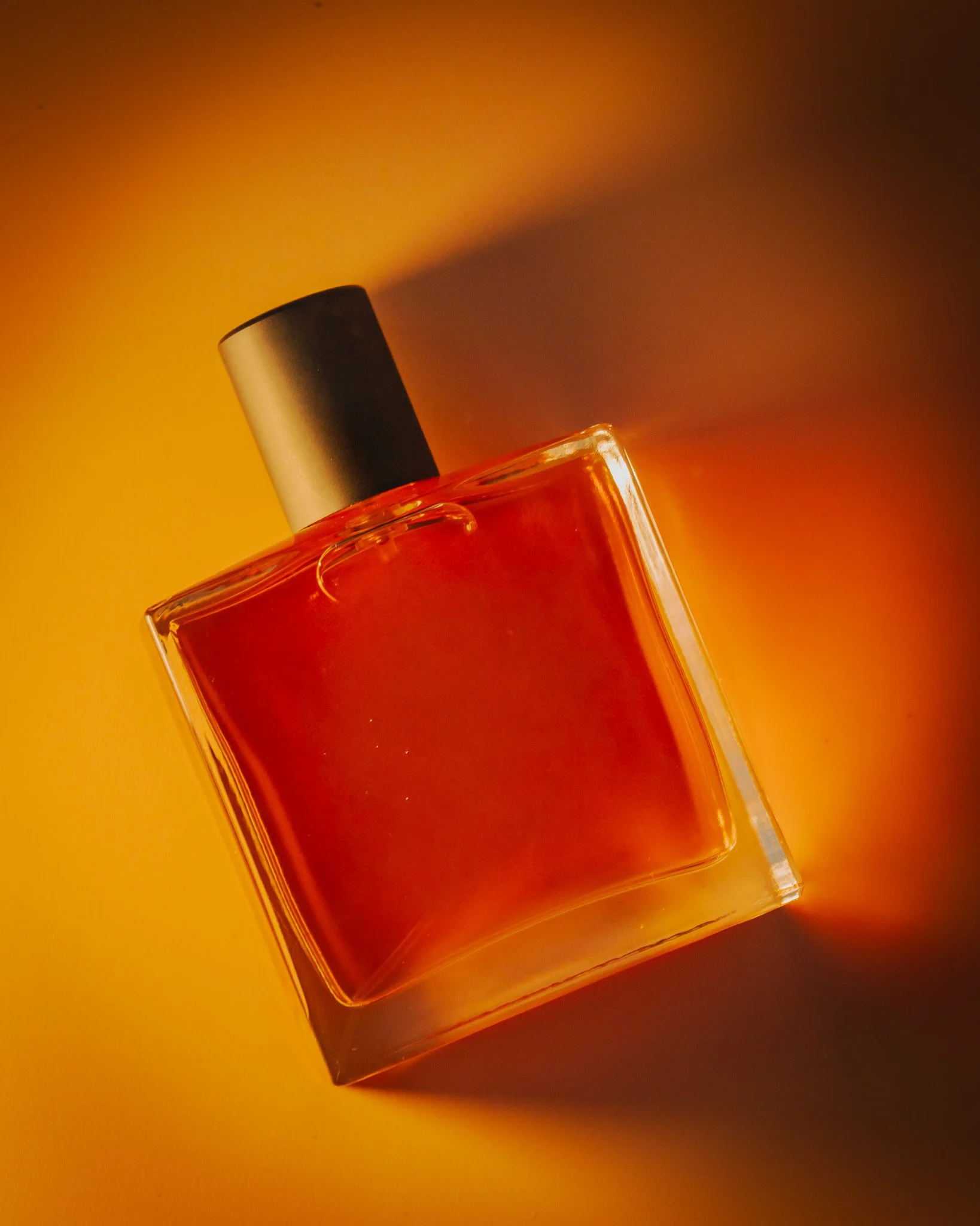 A square glass perfume bottle with a black cap on an orange background, casting a shadow.
