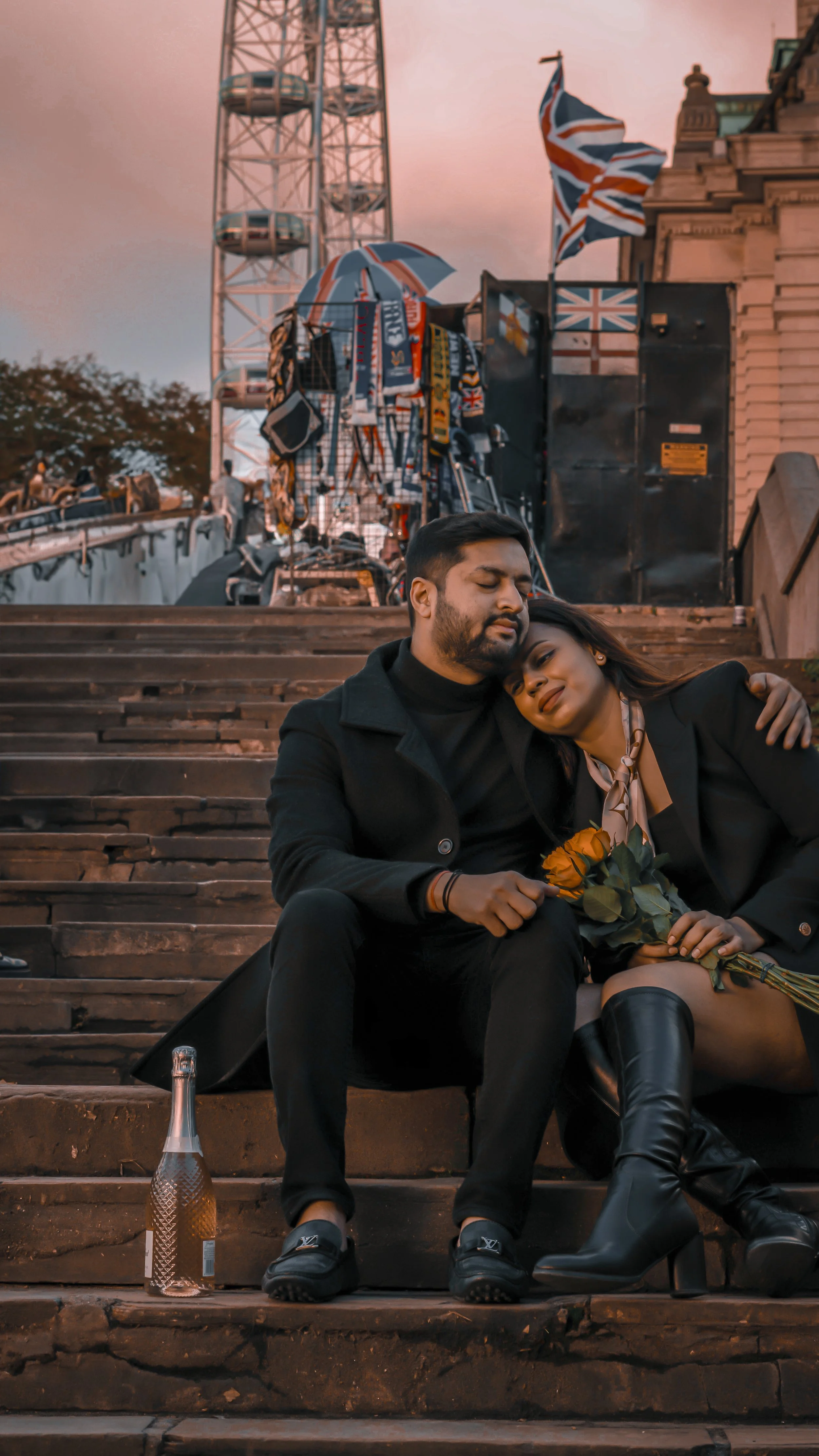 A couple sitting on stone steps at dusk, embracing with closed eyes, holding a rose and a bottle of champagne beside them, with a ferris wheel and Union Jack flags in the background.