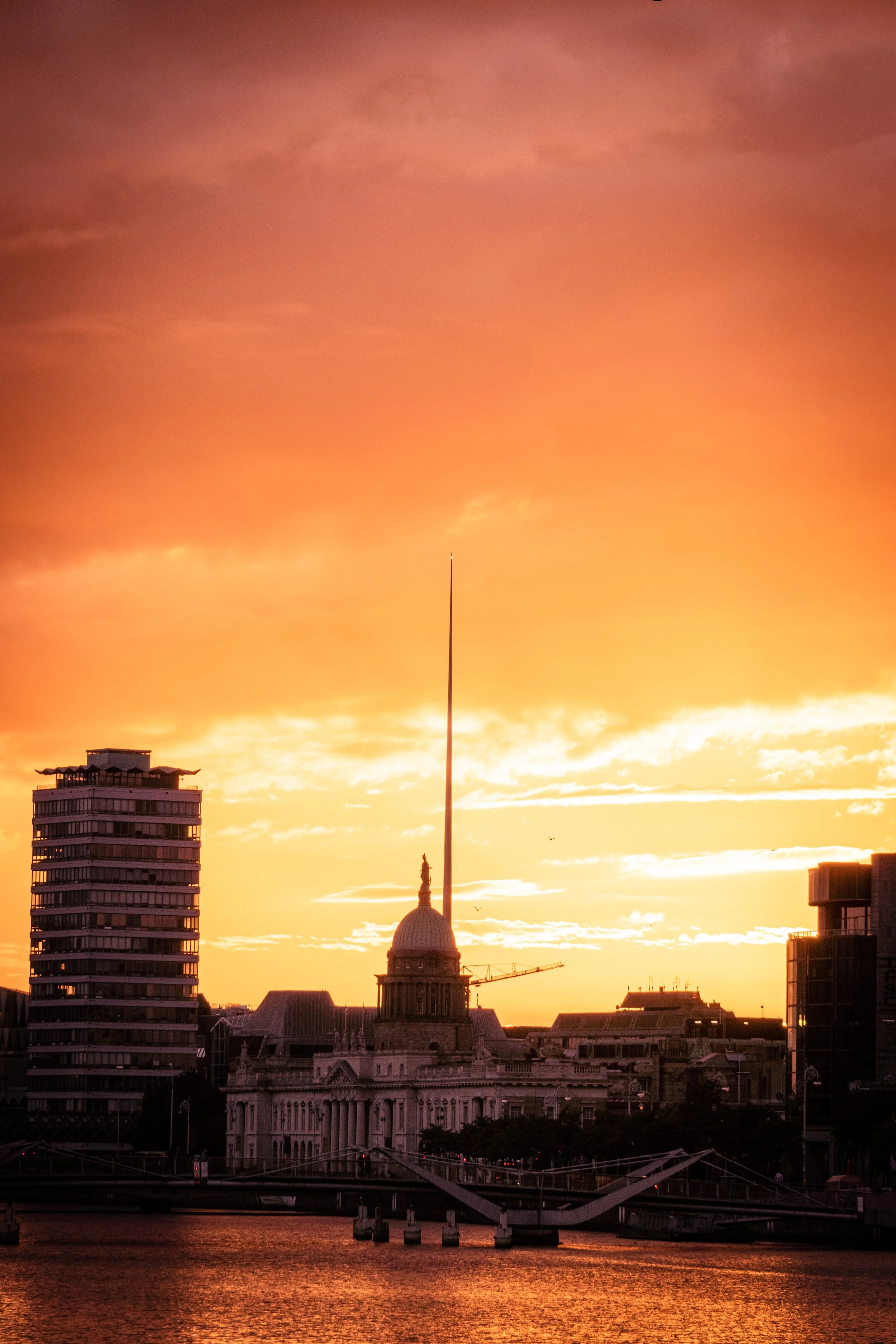 Sunset over a cityscape with a historic building featuring a dome and a tall flagpole, modern high-rise buildings, and a waterway in the foreground.