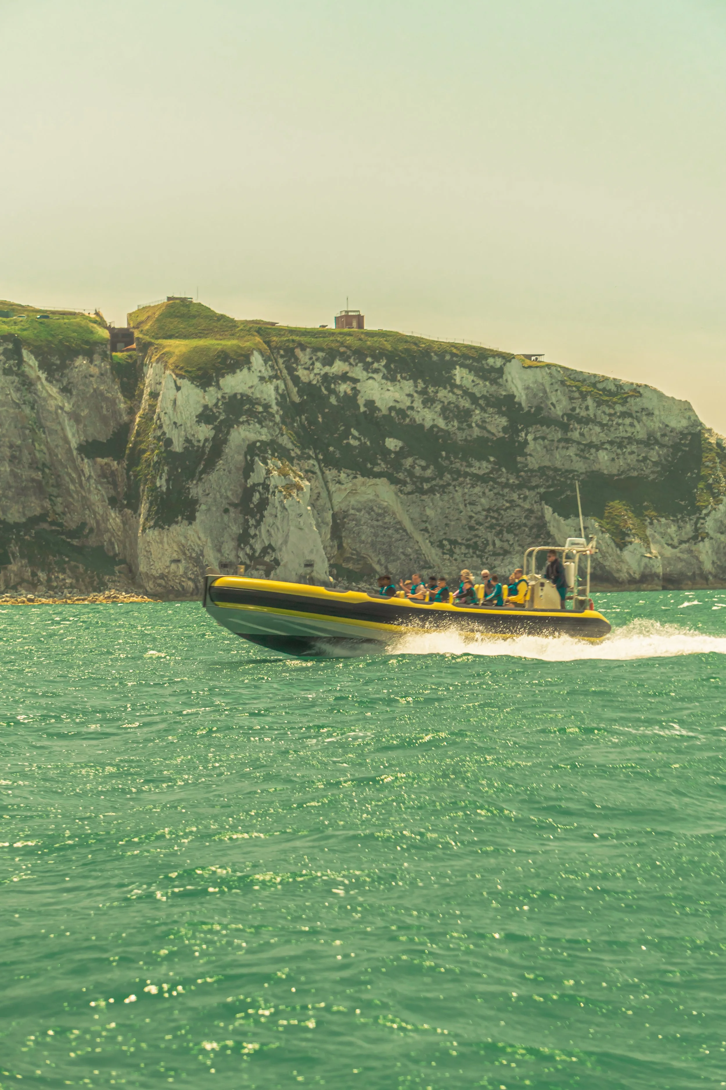 A speedboat with passengers traveling on green water near a rocky cliff with buildings on top.