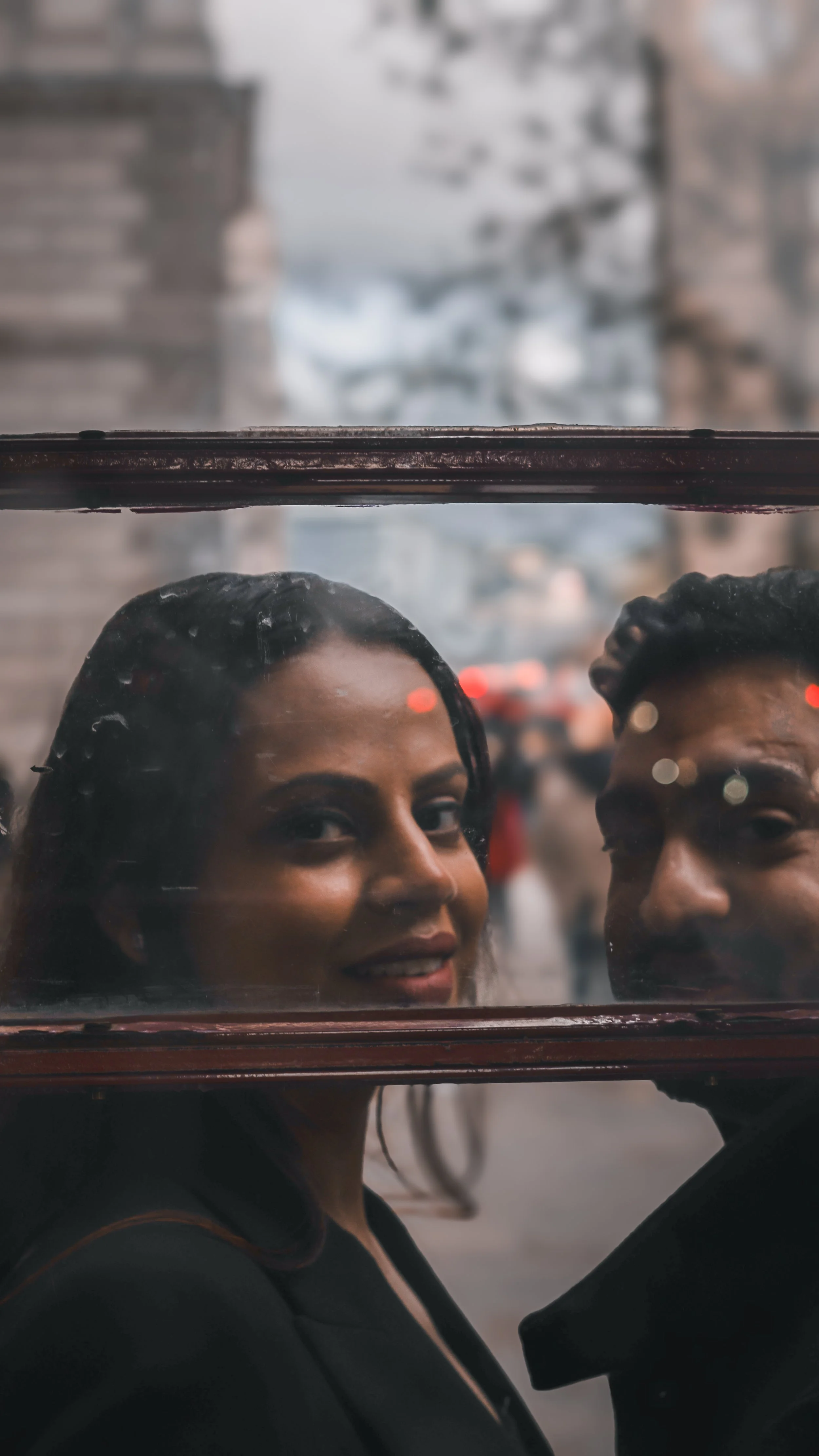 A woman with dark hair and a man with dark, curly hair are seen through a window with dirt and smudges. The woman is smiling and the background is blurred with city lights.