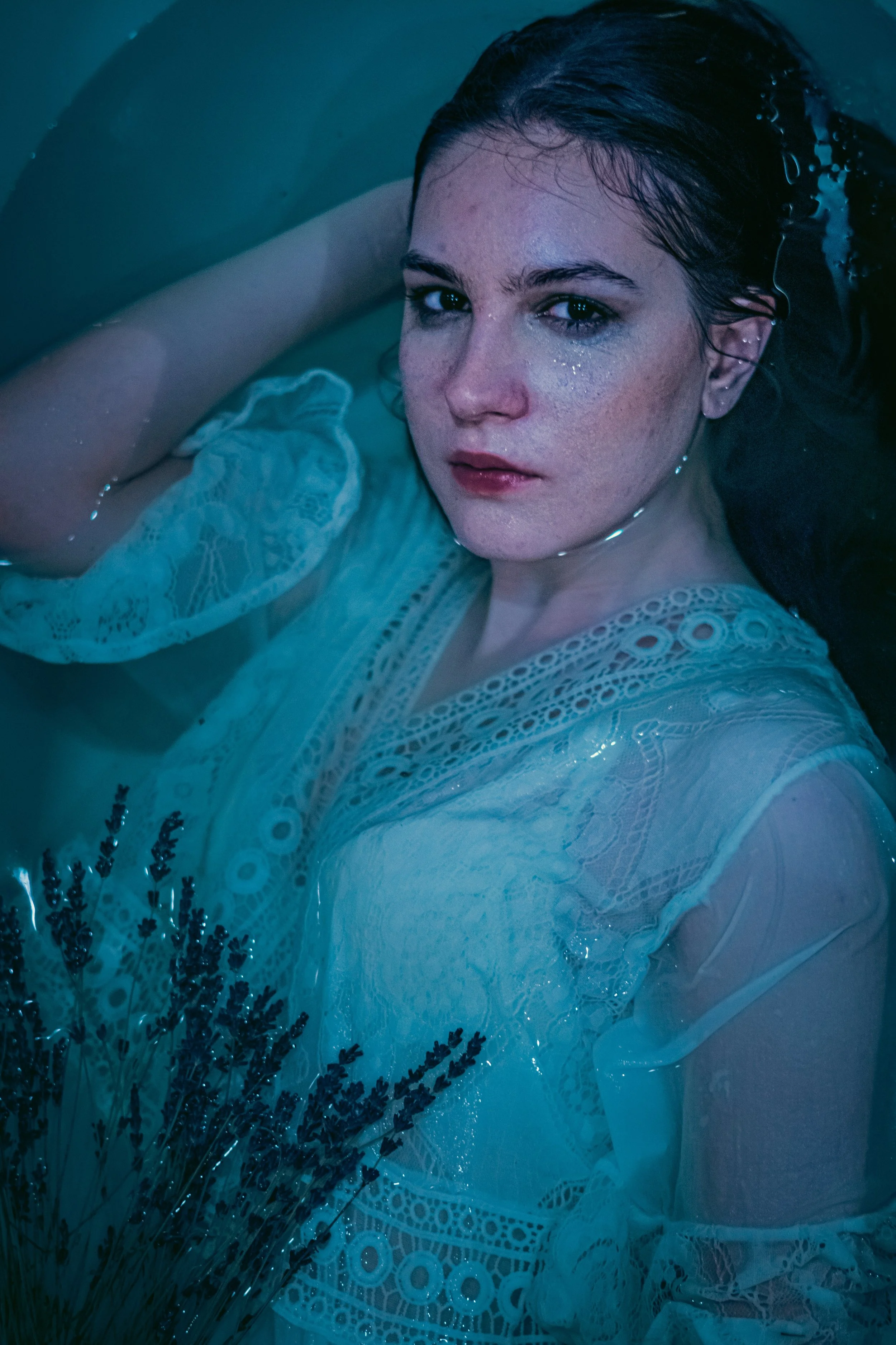 A woman with wet hair and a white lace dress lying in water, looking at the camera with a serious expression.