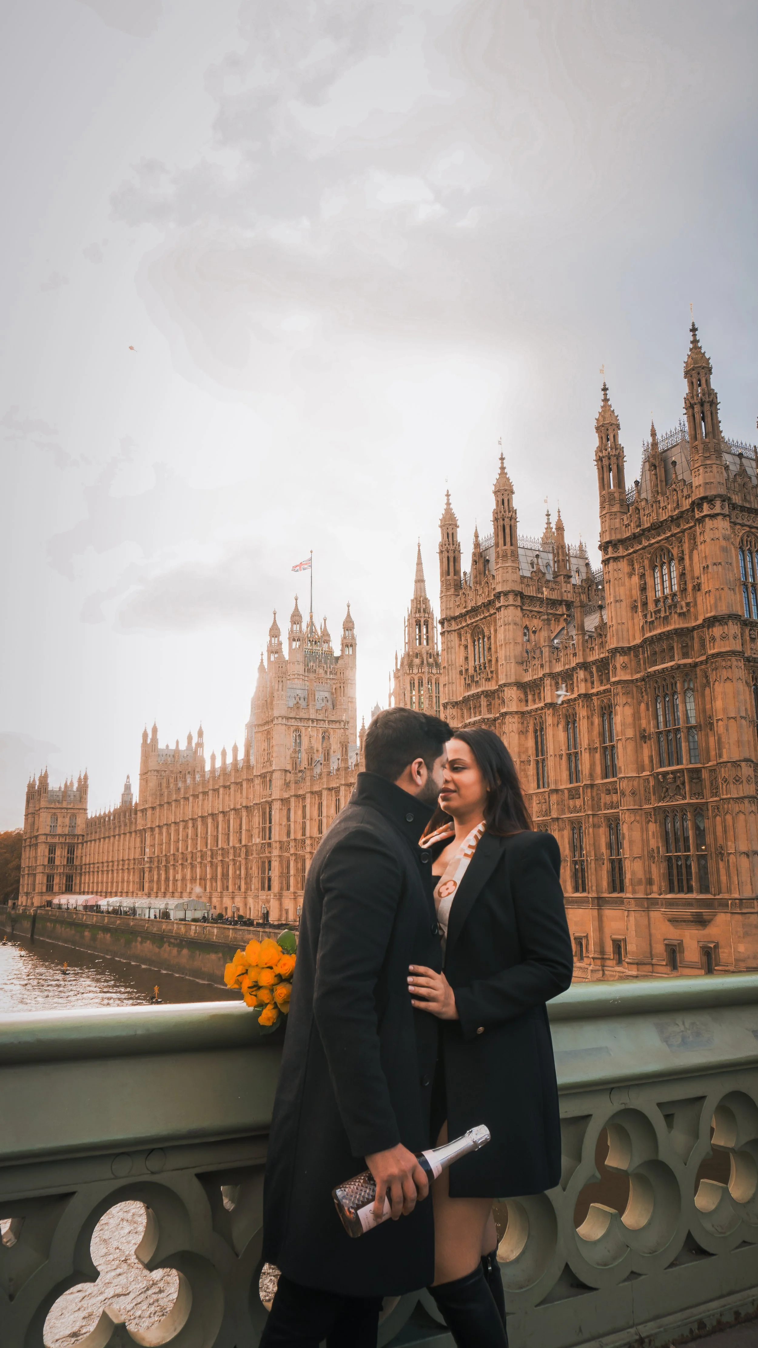 A couple standing close together on a bridge with the Houses of Parliament in London in the background, holding a bottle of champagne and a bouquet of yellow flowers, during daytime with cloudy skies.