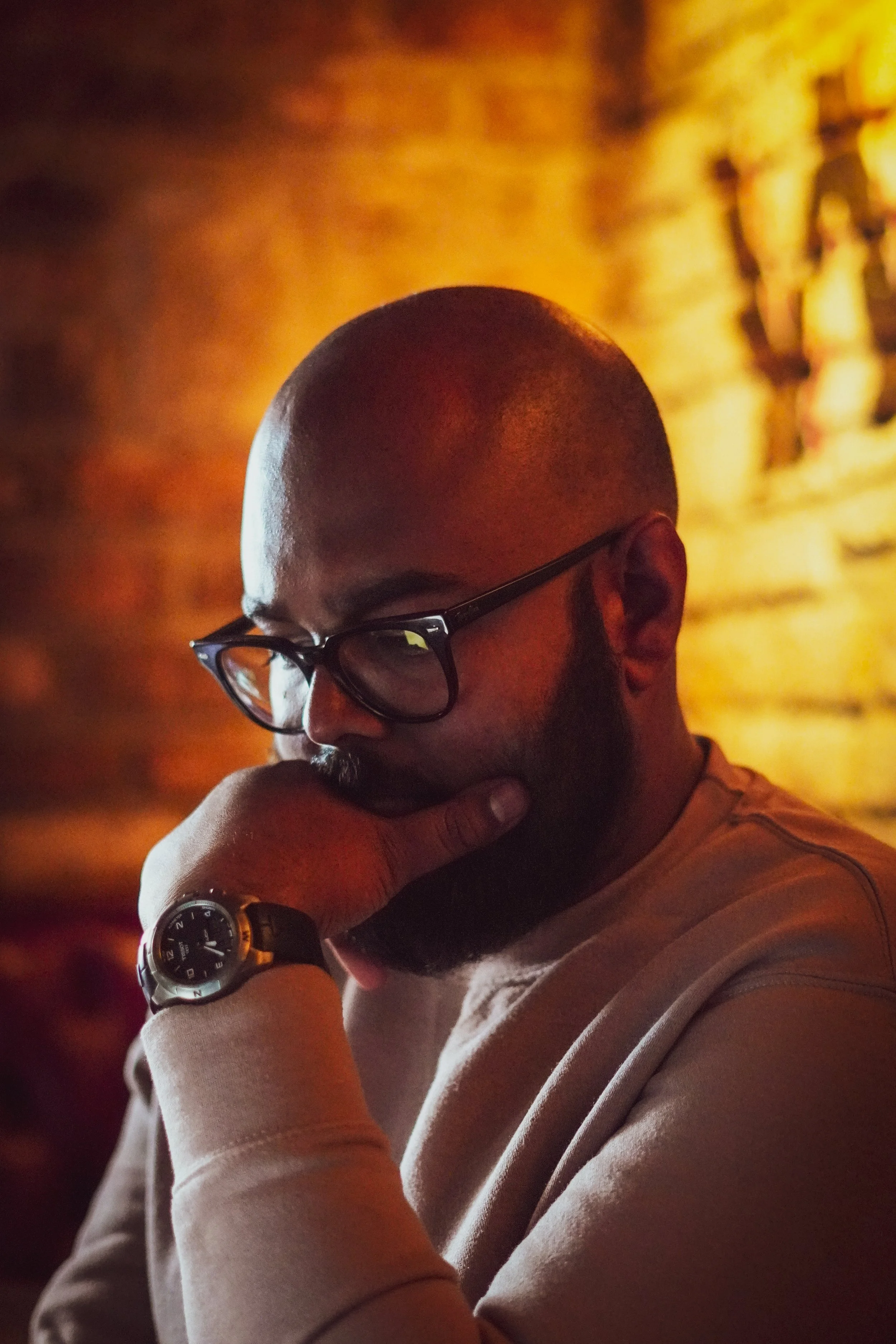 A man with a shaved head and glasses resting his chin on his hand in a dimly lit room with warm lighting.