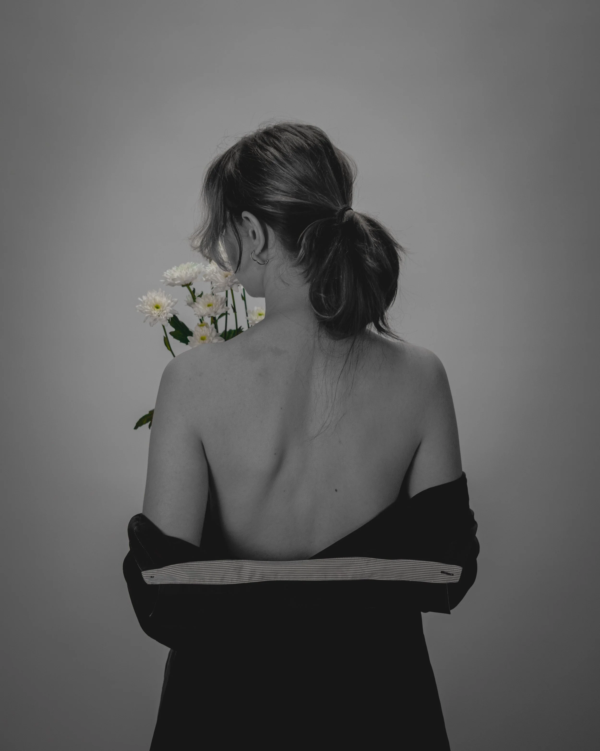 Black and white photo of a woman with her back to the camera, holding a bouquet of daisies, wearing a partially off-the-shoulder top.