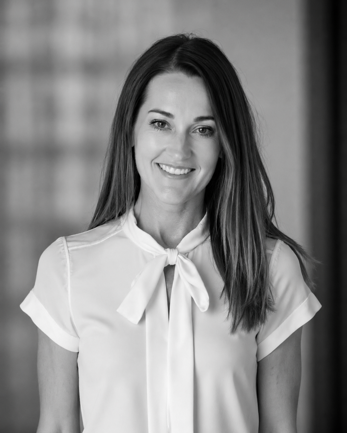 Black and white portrait of a smiling woman with long hair, wearing a blazer, in an office setting.