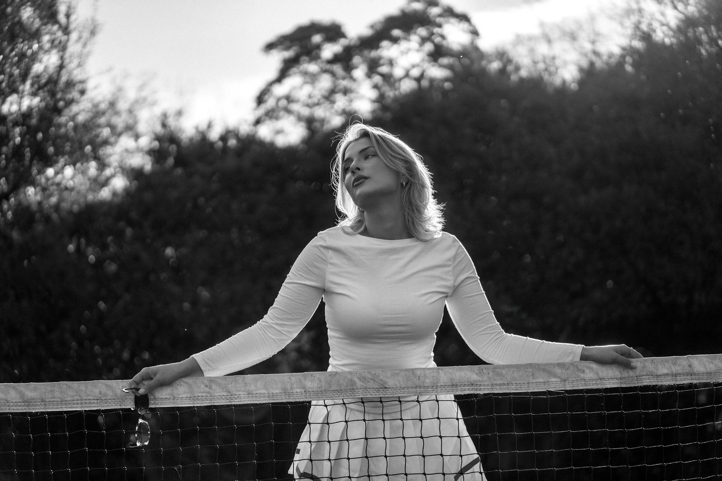 Melbourne model Lucille King, wearing tennis gear, back lit, stands in front of (holding) the tennis net. Image taken with Tamron 90mm F/2.8 Di III Macro VXD lens.