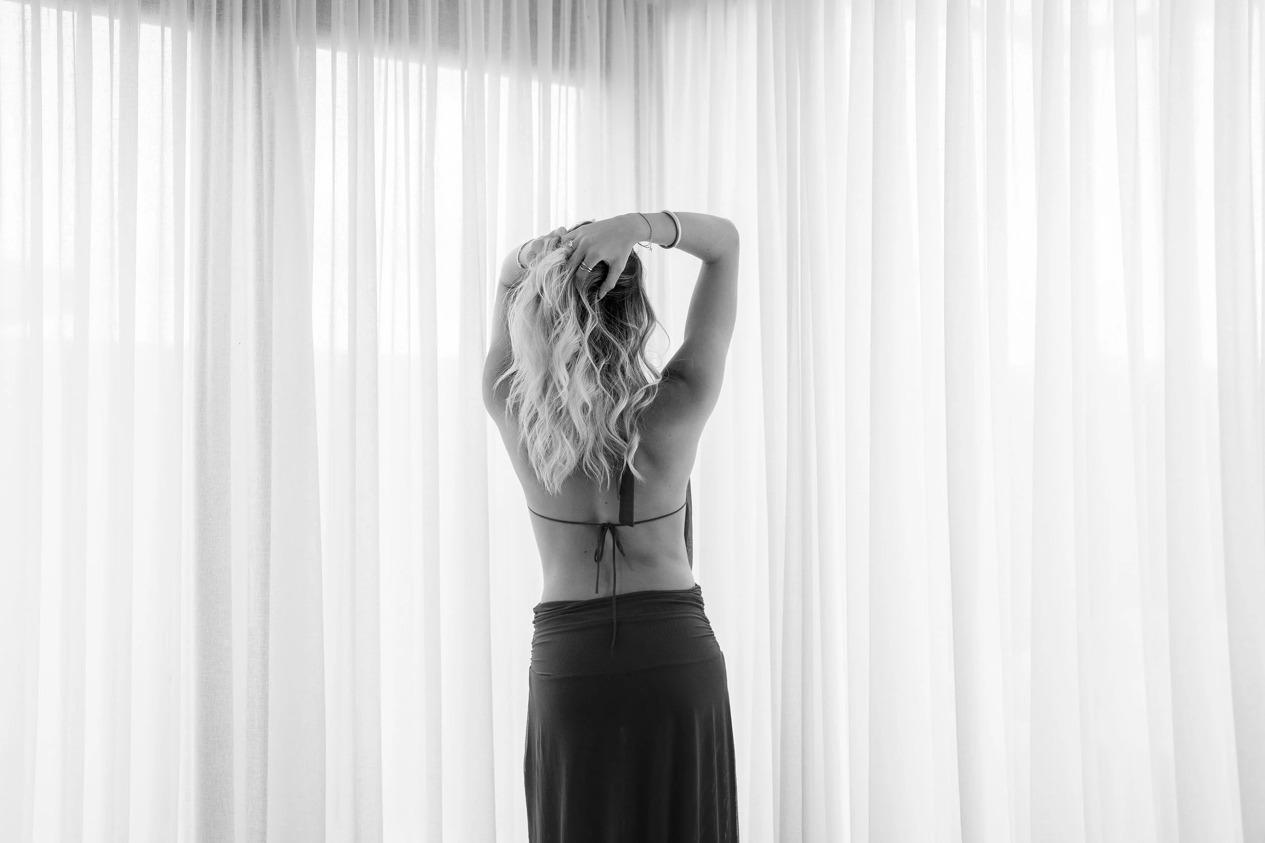 Model Isabella Duric, rear view, hands up in her blond hair, wearing an evening dress, standing in front of a curtain.
