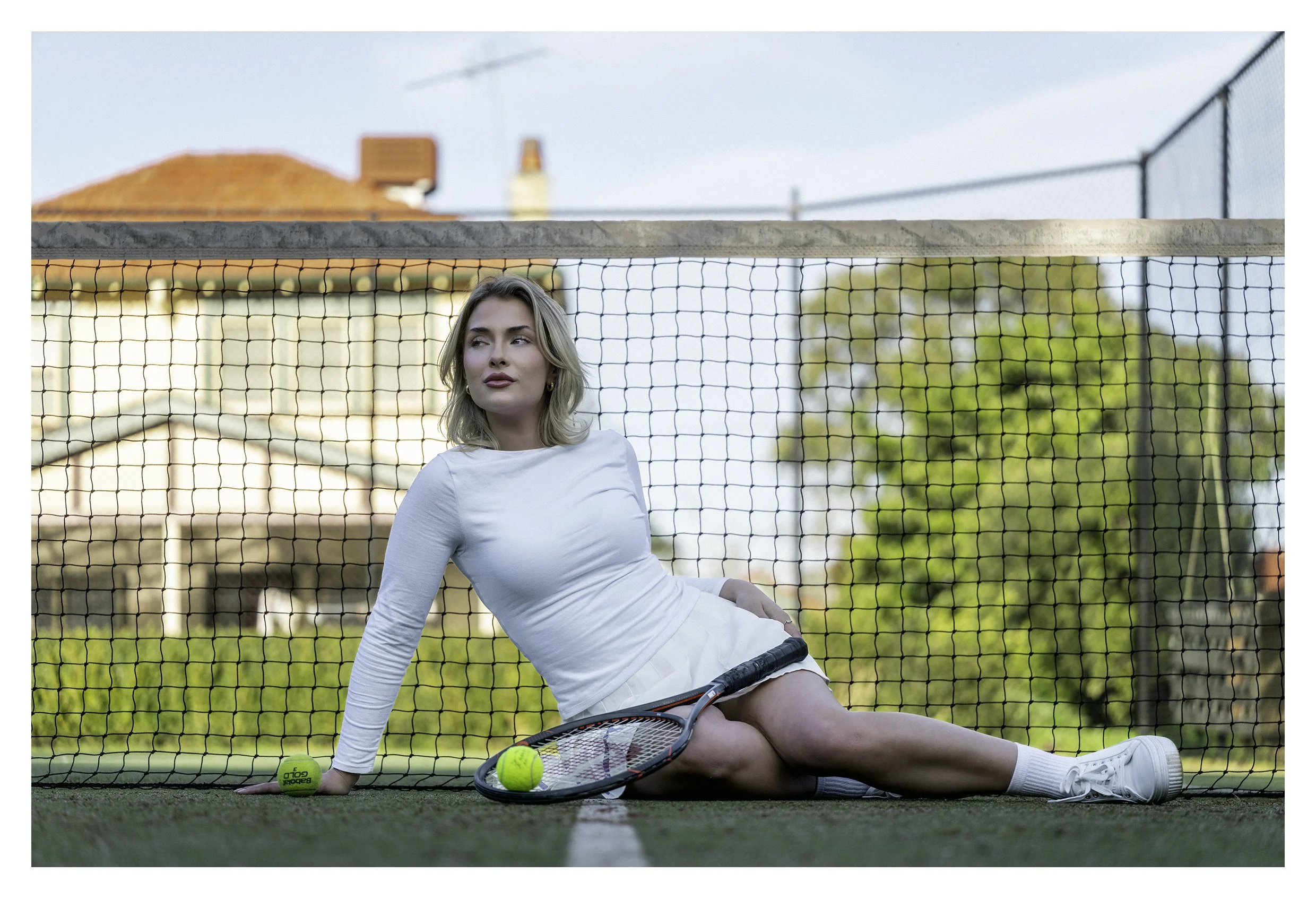 Model Lucille King rests, on the ground, of a tennis court, wearing white tennis clothes, a tennis racquet. Image taken with Tamron 90mm Macro lens.