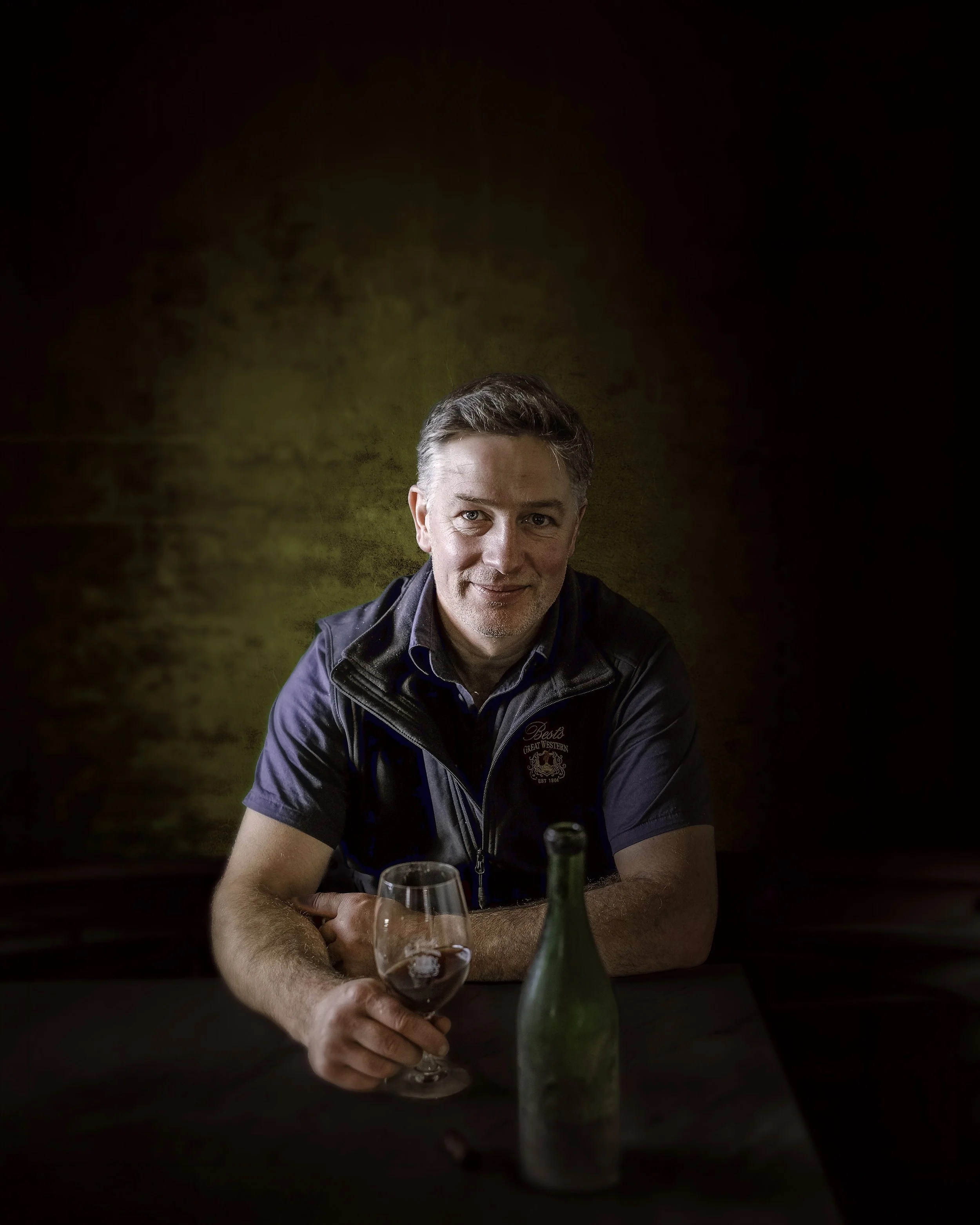 Winemaker Justin Purser sits at a table, wearing a Best's wines jacket, with a glass of Best's wine in front of him.