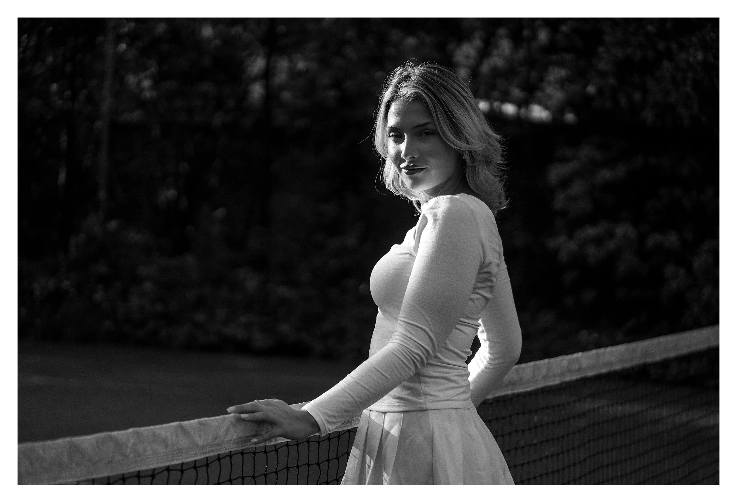Model Lucille King stands side-on to the camera, wearing tennis gear, on a tennis court, resting against the tennis net. Image taken with Tamron 90mm F/2.8 Di III Macro VXD lens.