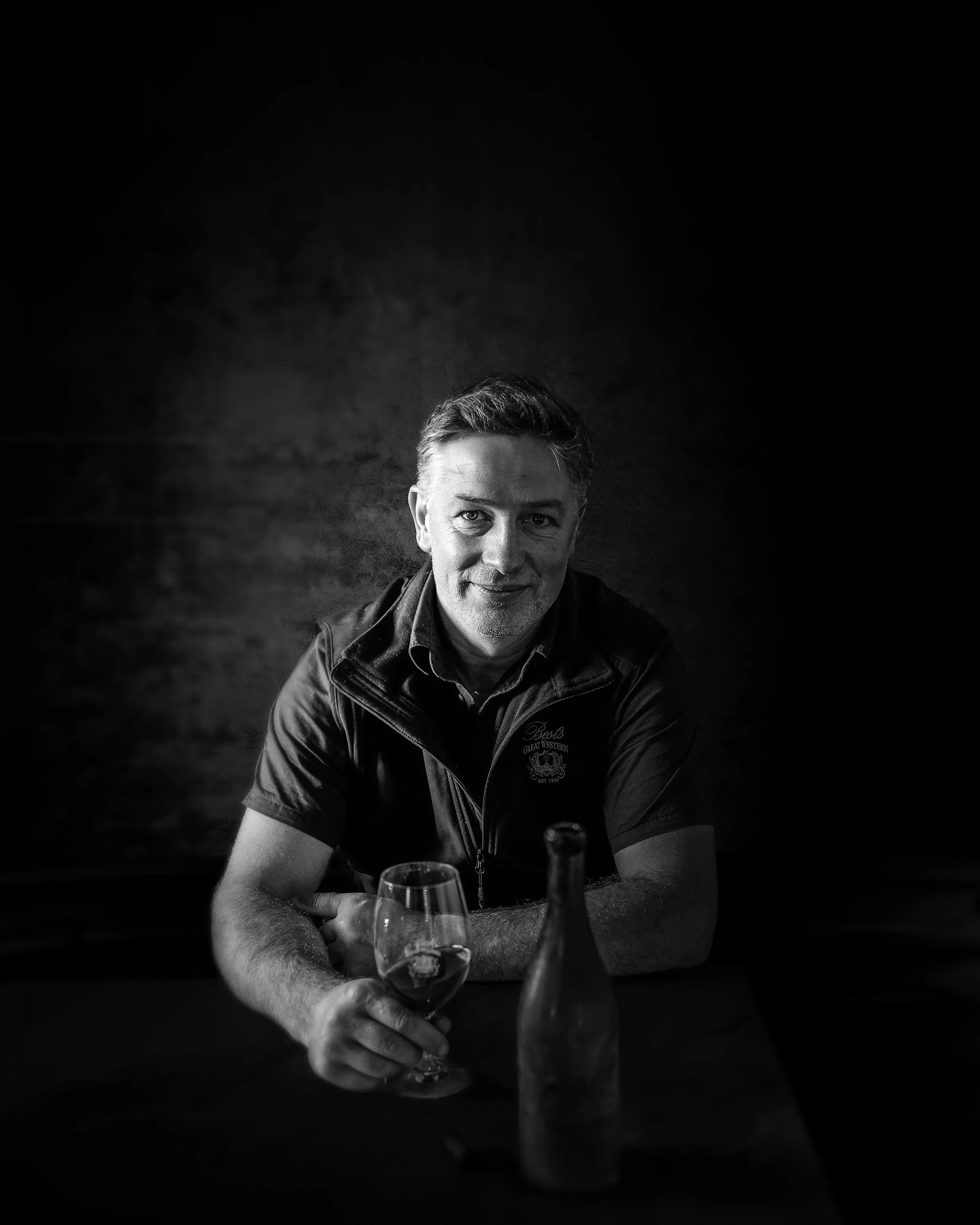 Black and white image of winemaker Justin Purser sits at a table, wearing a Best's wines jacket, with a glass of Best's wine in front of him.