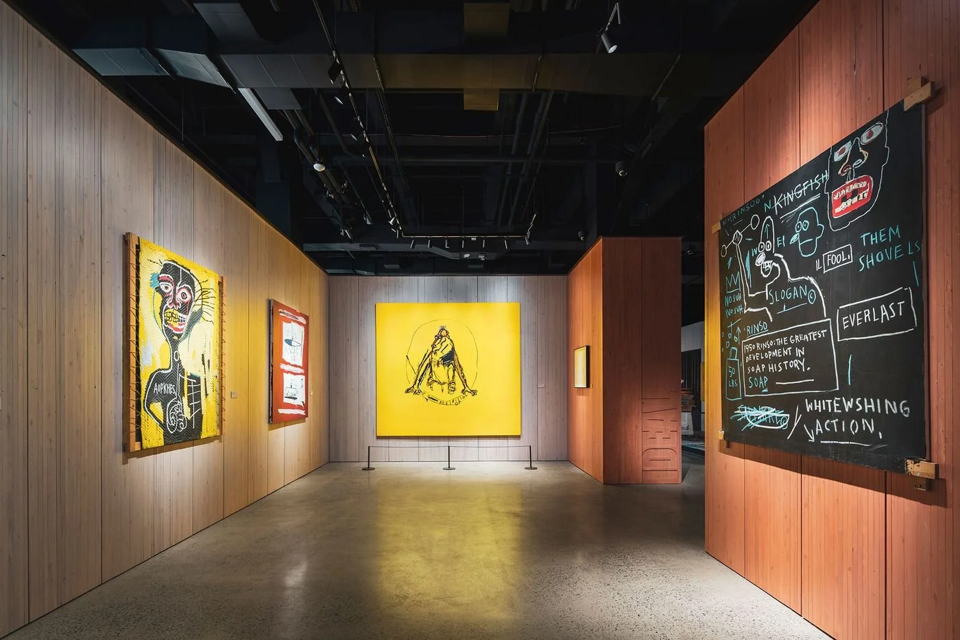 Art gallery with wood-paneled walls displaying colorful paintings, including a yellow background with a sketch of a person in the center, and a chalkboard with handwritten notes on the right.