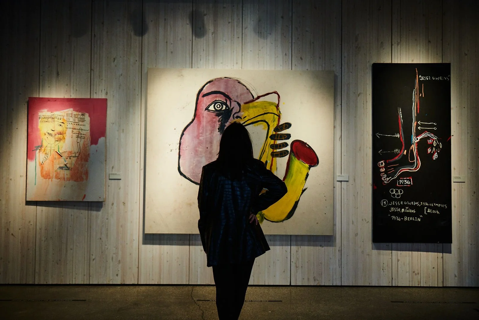 Person standing in front of three abstract art paintings in a gallery, one depicting a stylized face with a beak, another with sketches and text, and the third featuring a diagram and writing.