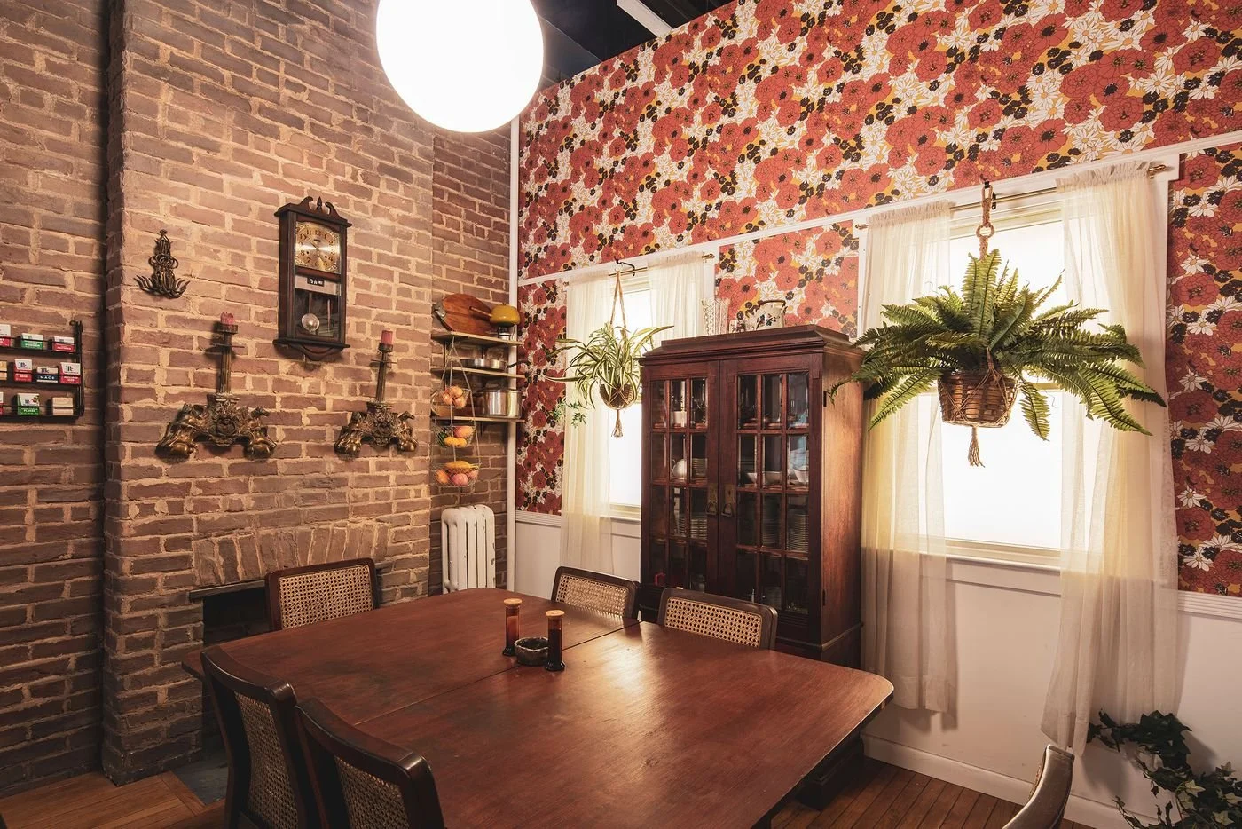 A dining room with a wooden table and six chairs, brick fireplace wall with antique wall sconces, a wooden clock, hanging plants, a glass cabinet with dishes, and floral wallpaper with two windows draped with sheer curtains.