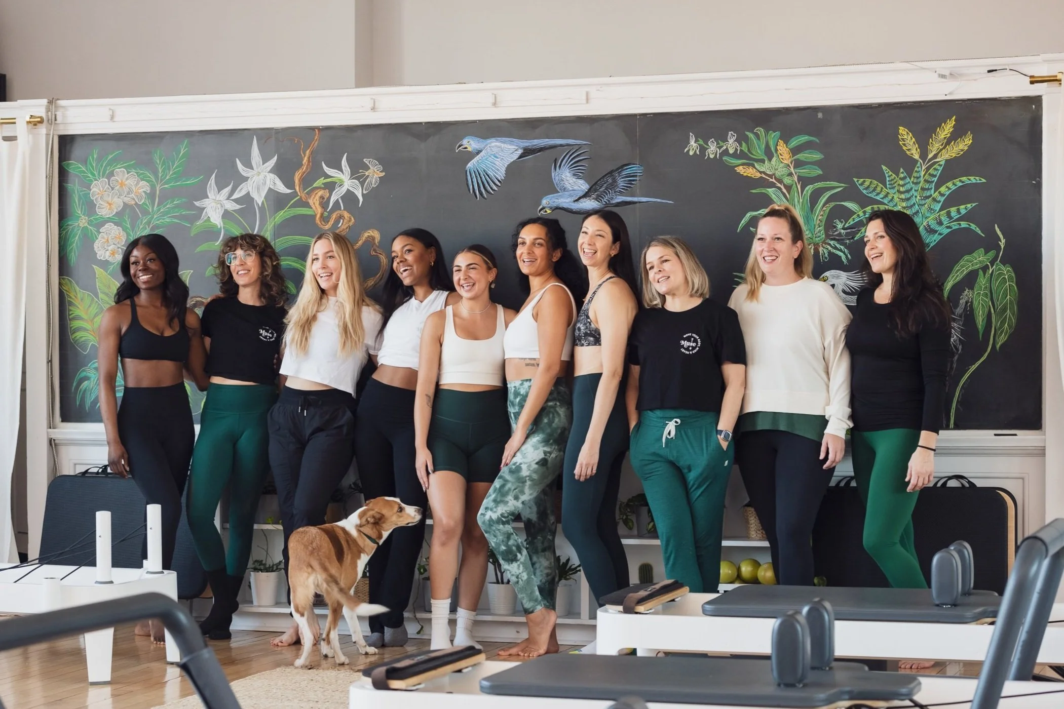 Group of Pilates Teachers standing together in a Pilates studio with a decorative blackboard featuring birds and plants in the background.