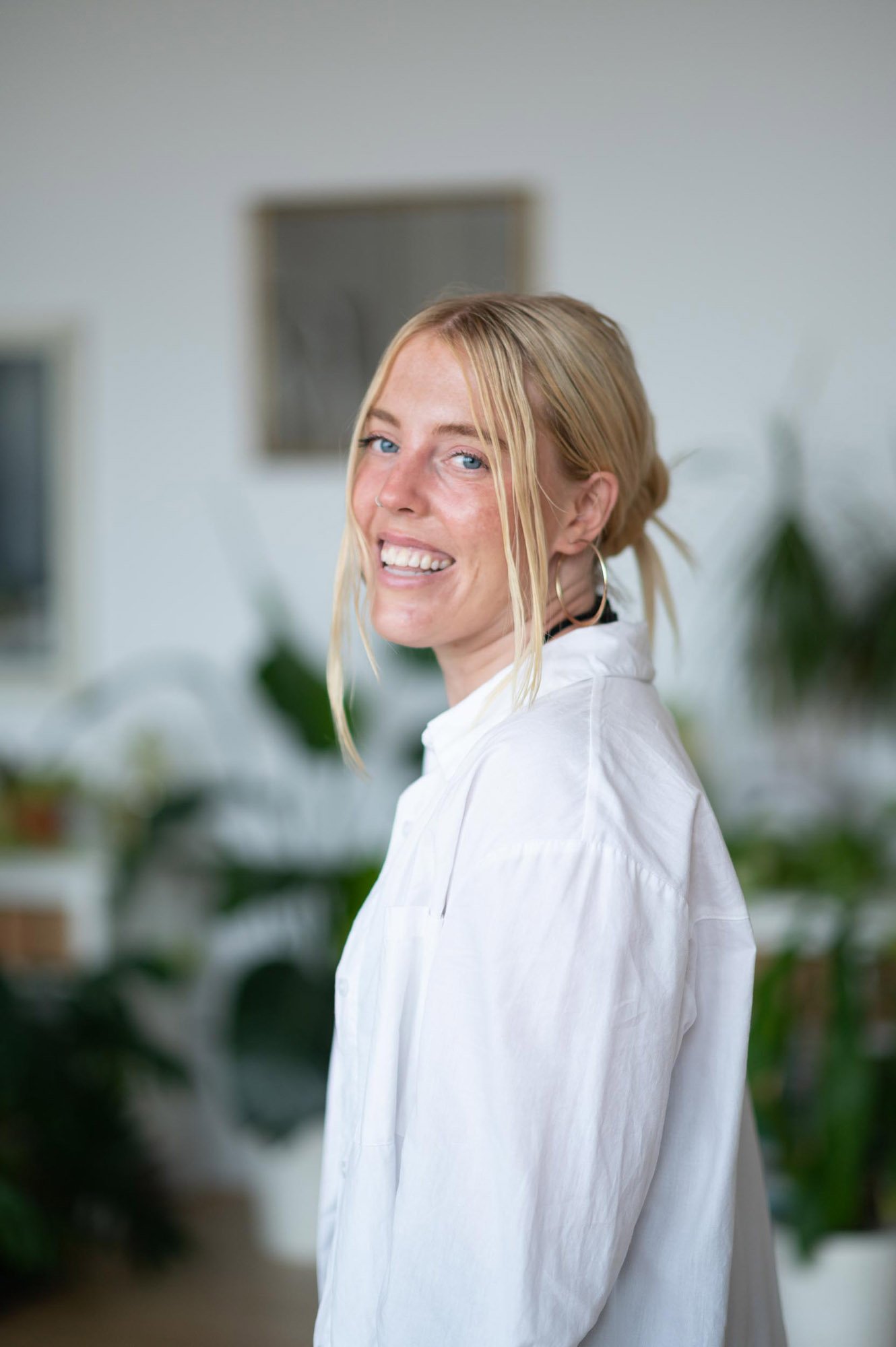 A Pilates Teacher Trainer woman with blonde hair, blue eyes, smiling and wearing a white shirt, in a Pilates studio filled with plants..